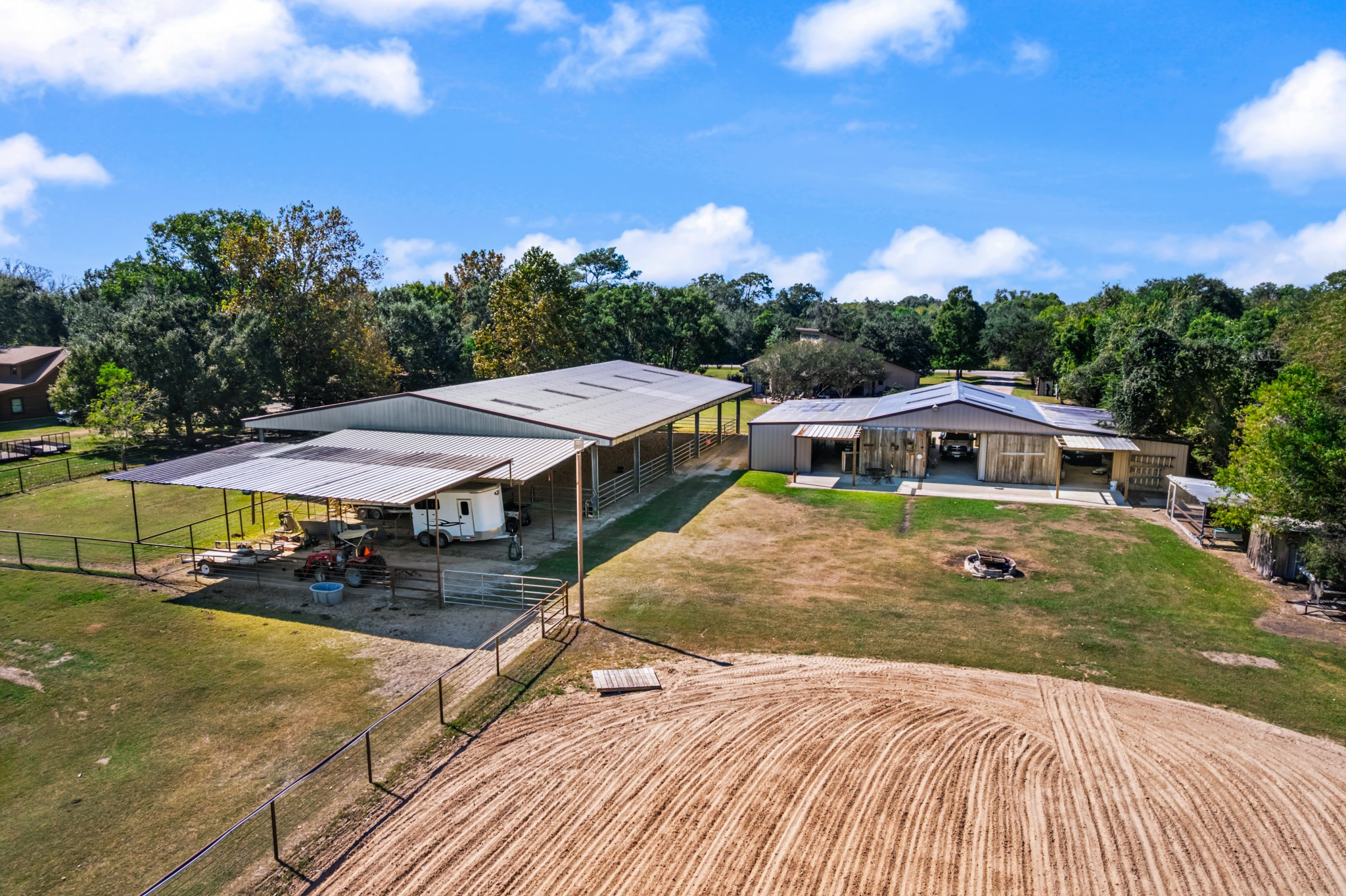 Aerial view highlighting the property's impressive equestrian facilities, including a spacious covered horse barn, fenced paddocks, covered trailer parking, and a large sand riding arena. An additional outbuilding provides versatile space for equipment, vehicles, or storage. Thoughtfully arranged for horse care, riding, and everyday ranch activities, this property offers an appealing setup for equestrian enthusiasts.
