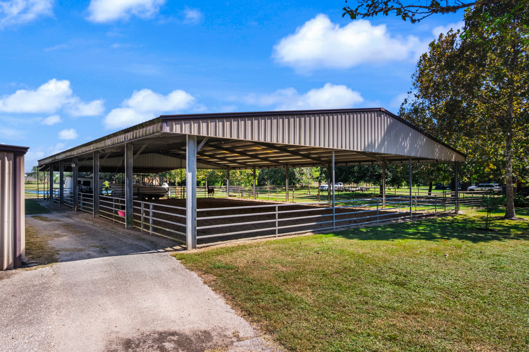 Impressive covered equestrian arena featuring a soaring metal roof, open sides for natural ventilation, and sturdy metal panel fencing. The spacious riding and training area offers protection from the elements, providing a versatile setup for working horses, practicing riding skills, and enjoying equestrian activities throughout the year.