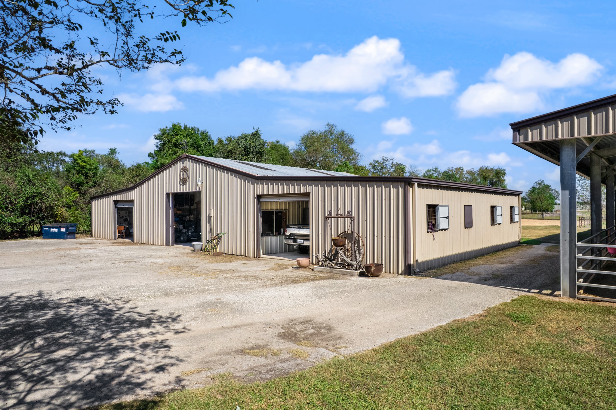 Expansive metal workshop and equestrian building featuring multiple overhead roll-up doors, dedicated work and storage areas, and an adjoining horse barn. Generous gravel parking and maneuvering space provide convenient access for vehicles, trailers, and equipment.