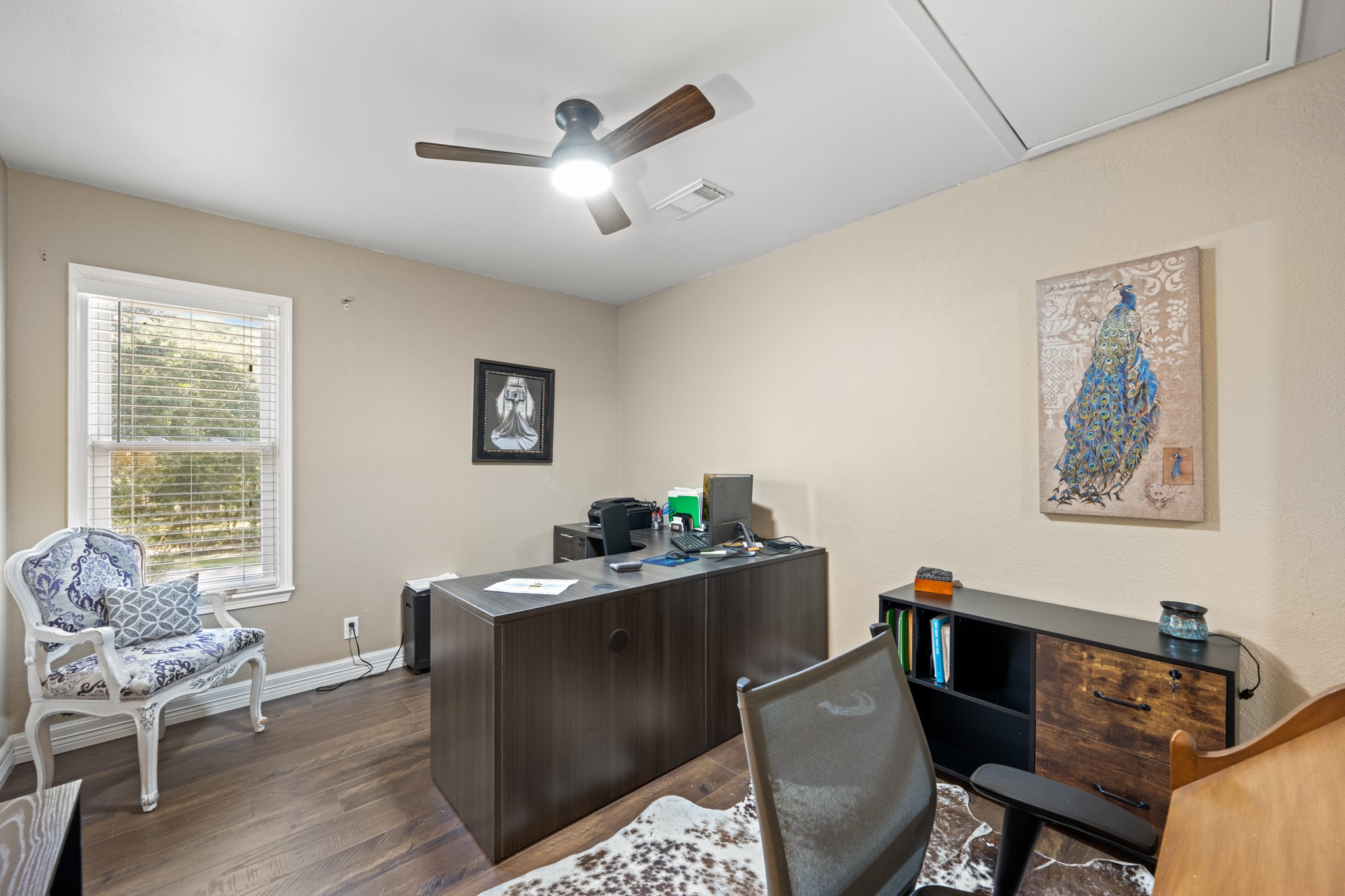 Versatile bedroom currently configured as a home office, featuring wood-look flooring, neutral tones, a ceiling fan, and a window providing natural light. Easily adaptable as an additional bedroom, study, or creative workspace to suit your needs.