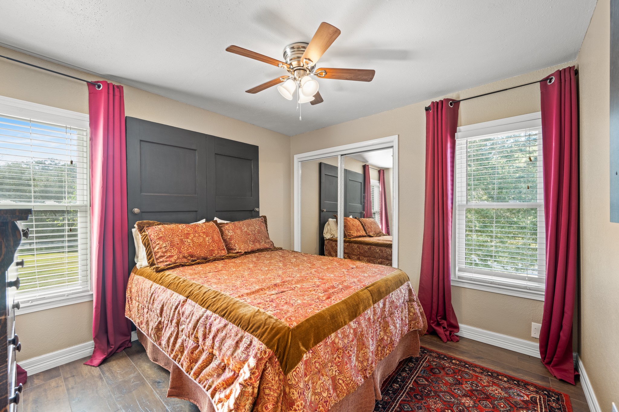 Bright secondary bedroom features windows on two walls, mirrored closet doors, wood-look flooring, and a ceiling fan, creating a comfortable and functional retreat.