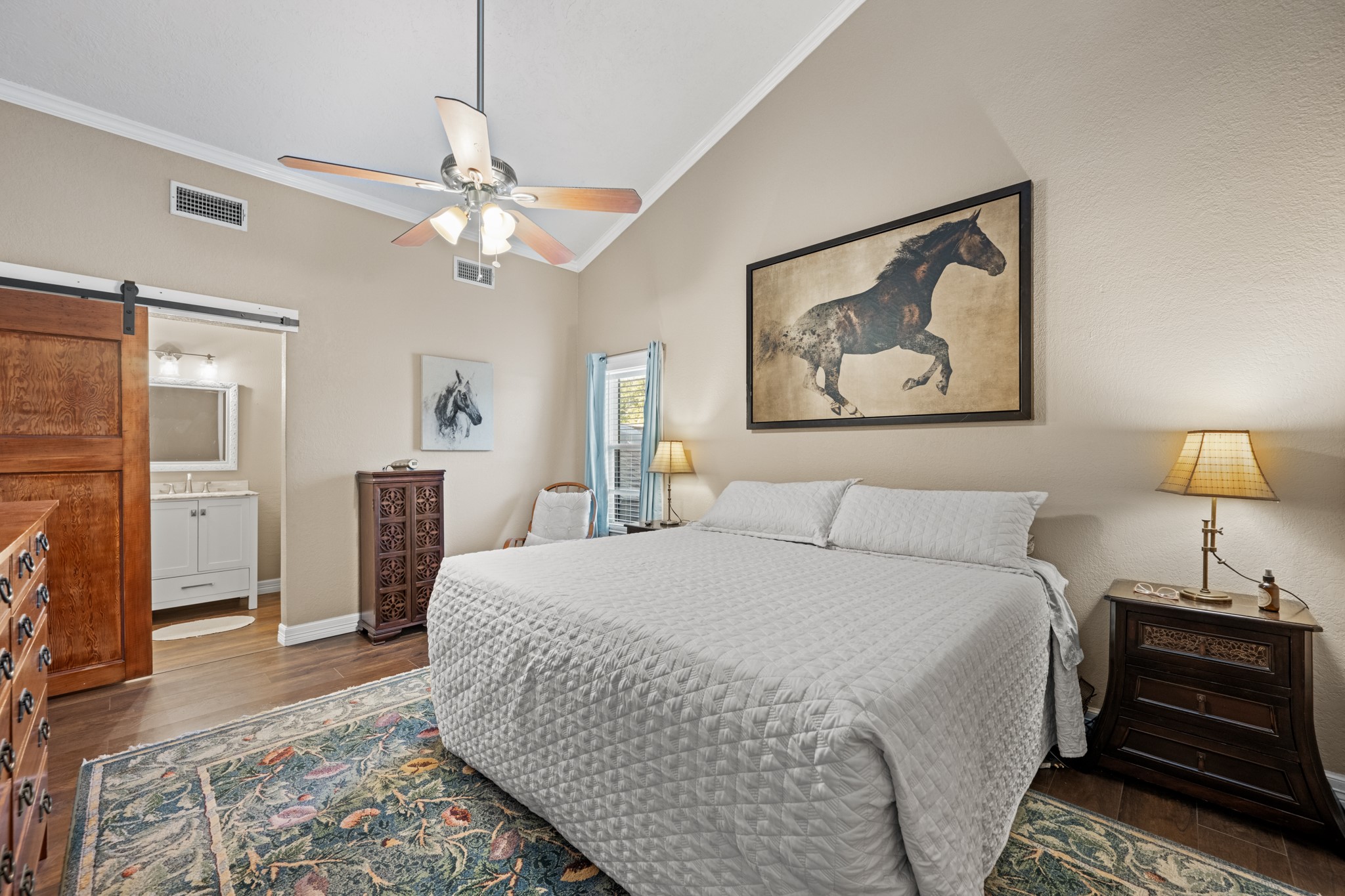 Spacious primary suite features a vaulted ceiling, warm wood-look flooring, ceiling fan, and a rustic sliding barn door leading to the ensuite bathroom.