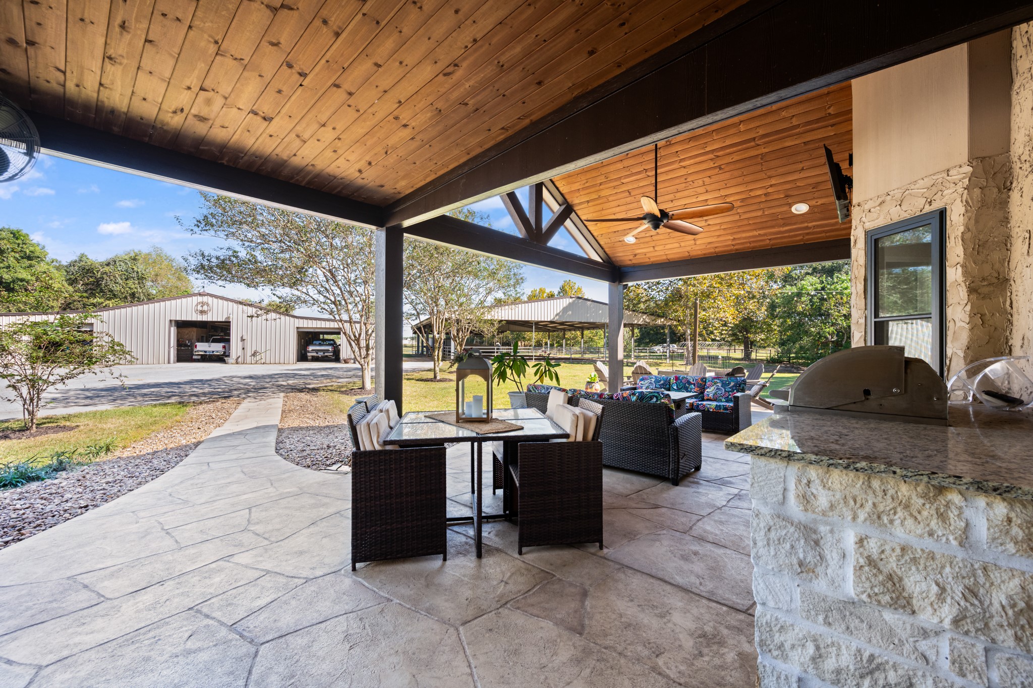 A second perspective highlights the covered outdoor kitchen with built-in stainless steel grill, stone-accented countertops, dining space, and comfortable lounge seating. The wood-plank ceiling and exposed timber beams add rustic elegance, with convenient access to the property's spacious outbuildings.