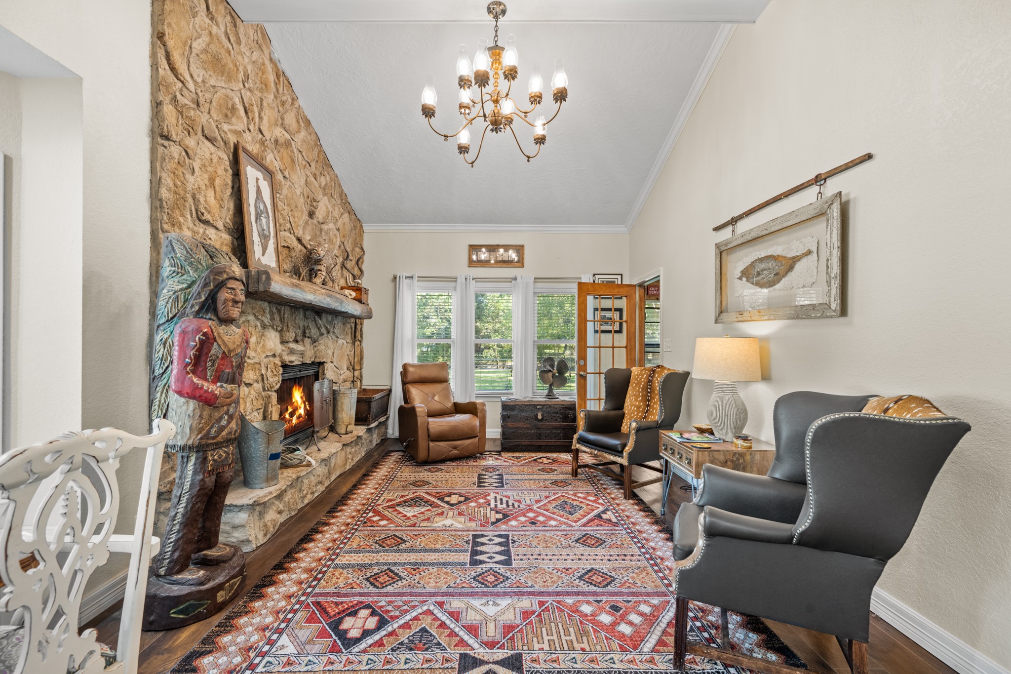 A dramatic floor-to-ceiling stone fireplace anchors this inviting living room, complemented by soaring ceilings, a rustic wood mantel, and large windows overlooking the property.