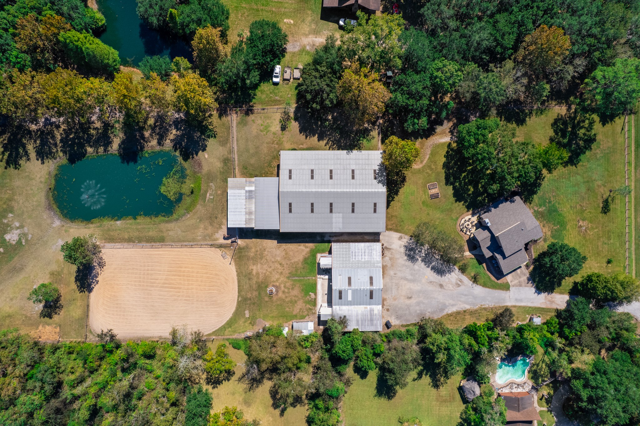 Overhead aerial view showcasing the property's impressive layout, featuring a spacious horse barn, sand riding arena, scenic pond with fountain, additional outbuilding, and residence surrounded by mature trees. The thoughtfully arranged equestrian facilities offer convenient access between the barn, arena, and surrounding grounds, creating an ideal setting for horse enthusiasts seeking space, functionality, and country living. Overhead aerial view showcasing the property's impressive layout, featuring a spacious horse barn, sand riding arena, scenic pond with fountain, additional outbuilding, and residence surrounded by mature trees. The thoughtfully arranged equestrian facilities offer convenient access between the barn, arena, and surrounding grounds, creating an ideal setting for horse enthusiasts seeking space, functionality, and country living.