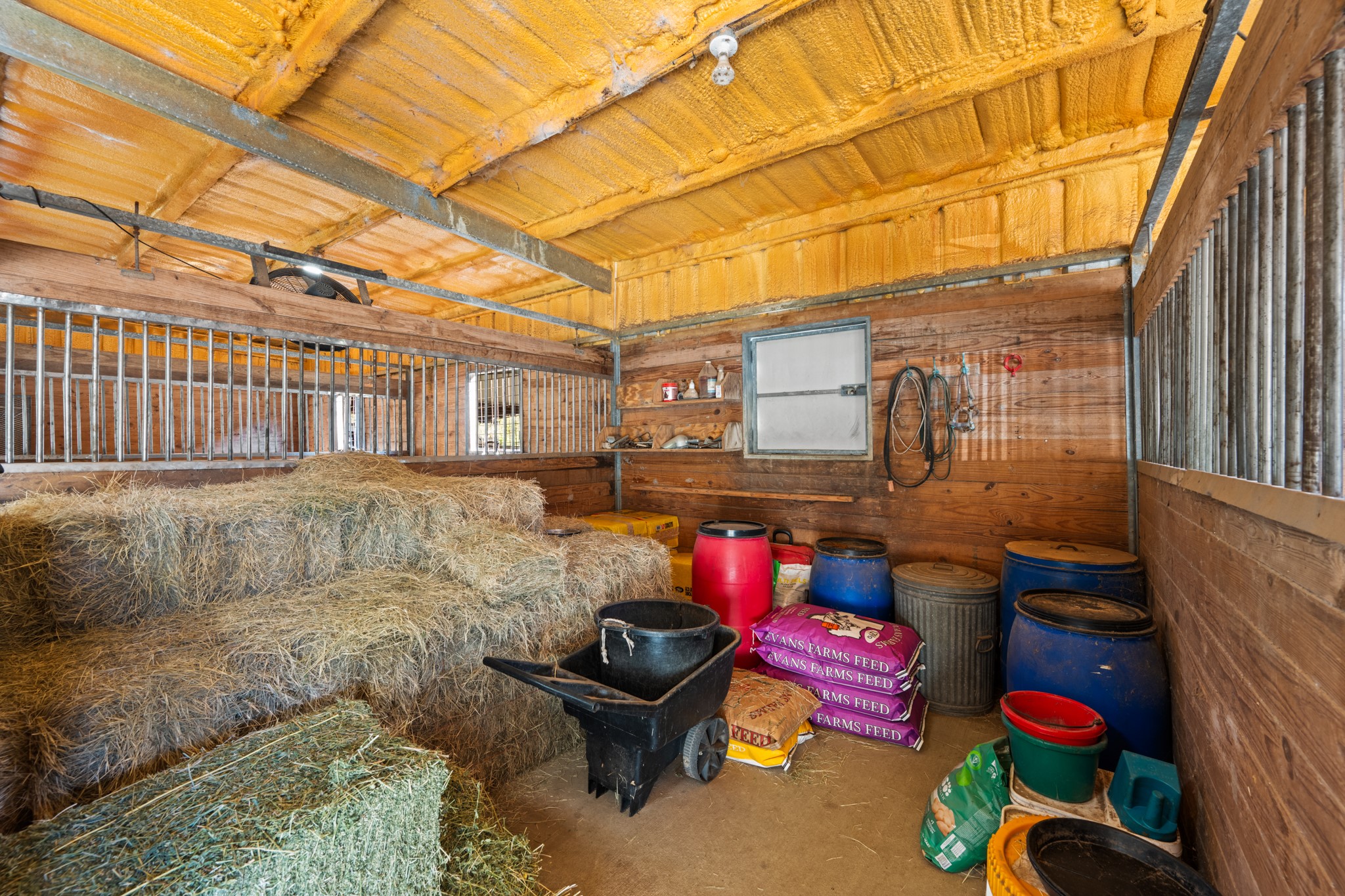 Dedicated feed and hay storage room/extra stall offers space for hay bales, feed supplies, and tack essentials, with wood-paneled walls, metal-bar partitions, and overhead insulation.