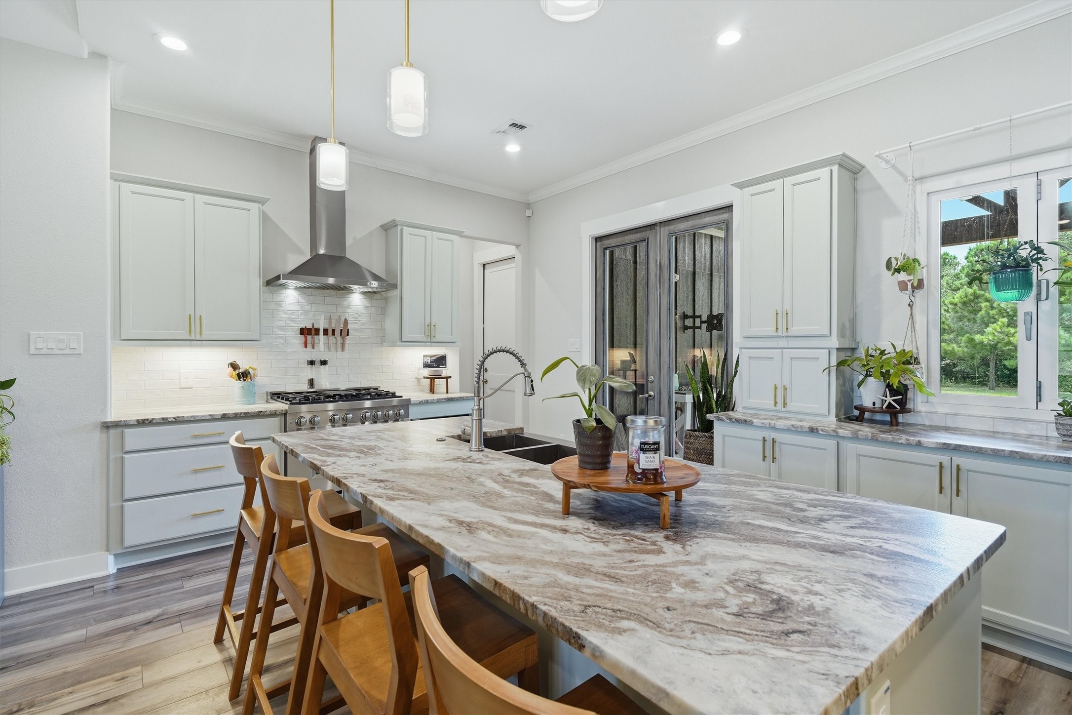 Large central island featuring leathered granite and pendant lighting. Stainless steel hood vent plus 6 burner range cooktop. Accordion style windows open to patio.