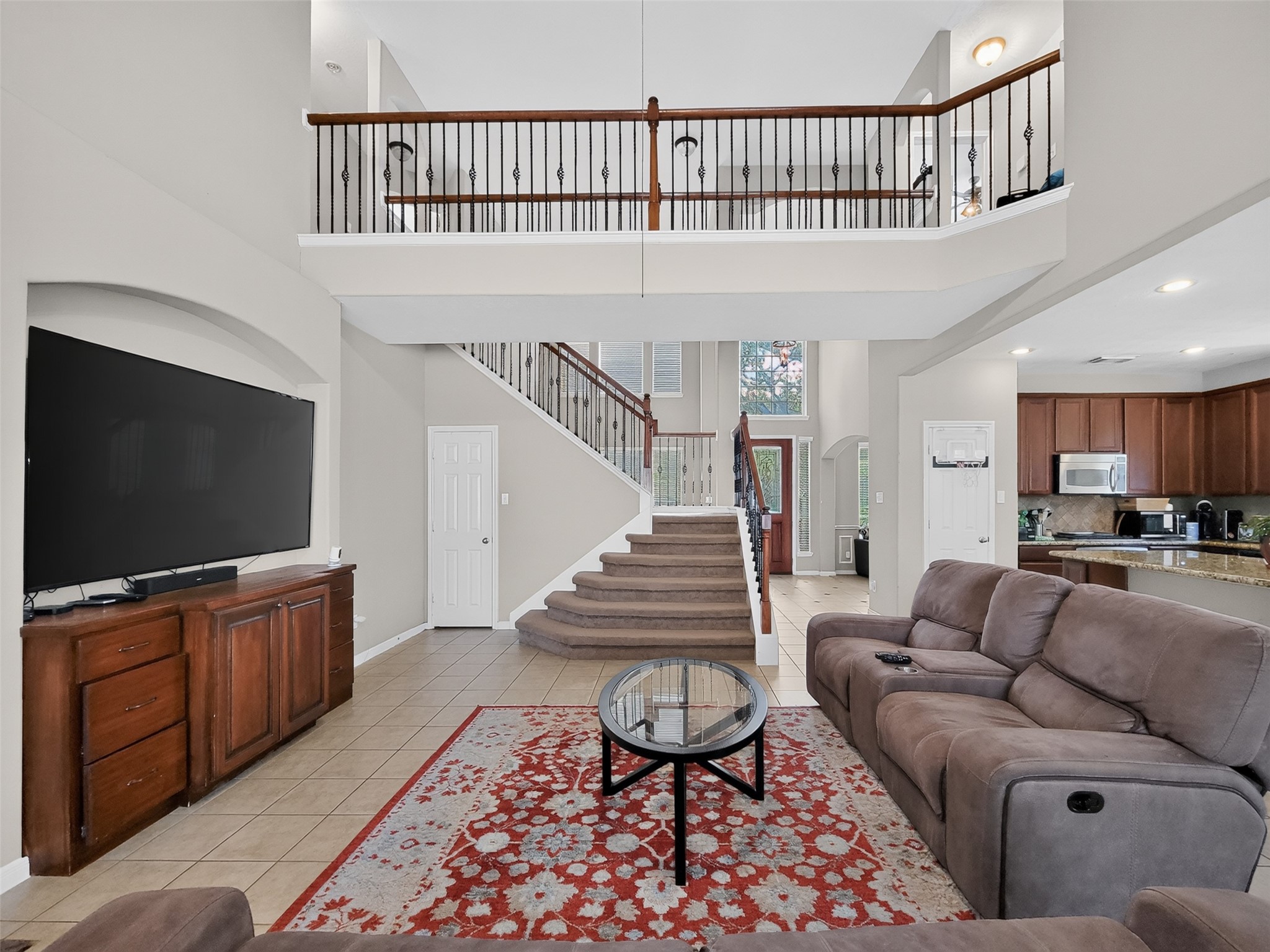 Spacious living room with high ceilings, a grand staircase, and an open view to the kitchen. Features a cozy seating area, large TV, and elegant railing on the upper level.