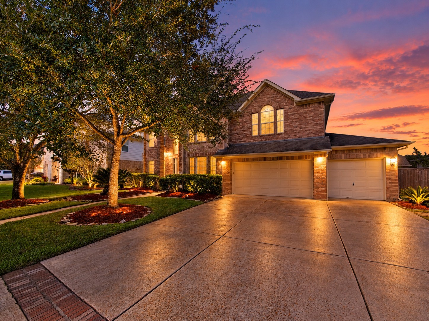 Stunning two-story brick home with a spacious three-car garage, beautifully lit at sunset, surrounded by lush landscaping and mature trees.