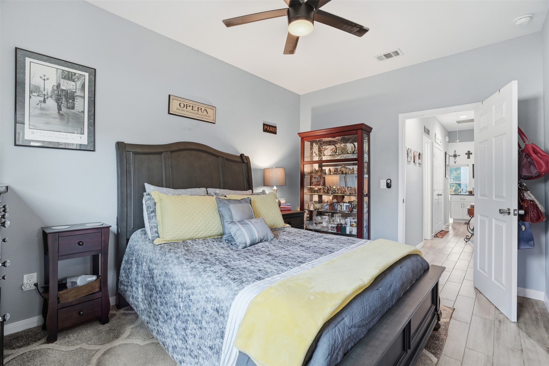 Primary bedroom with a stylish dark wood bed frame, complemented by soft gray walls and bright yellow accents. A glass display cabinet adds charm, while the ceiling fan ensures comfort. The room connects to a bathroom, enhancing convenience.