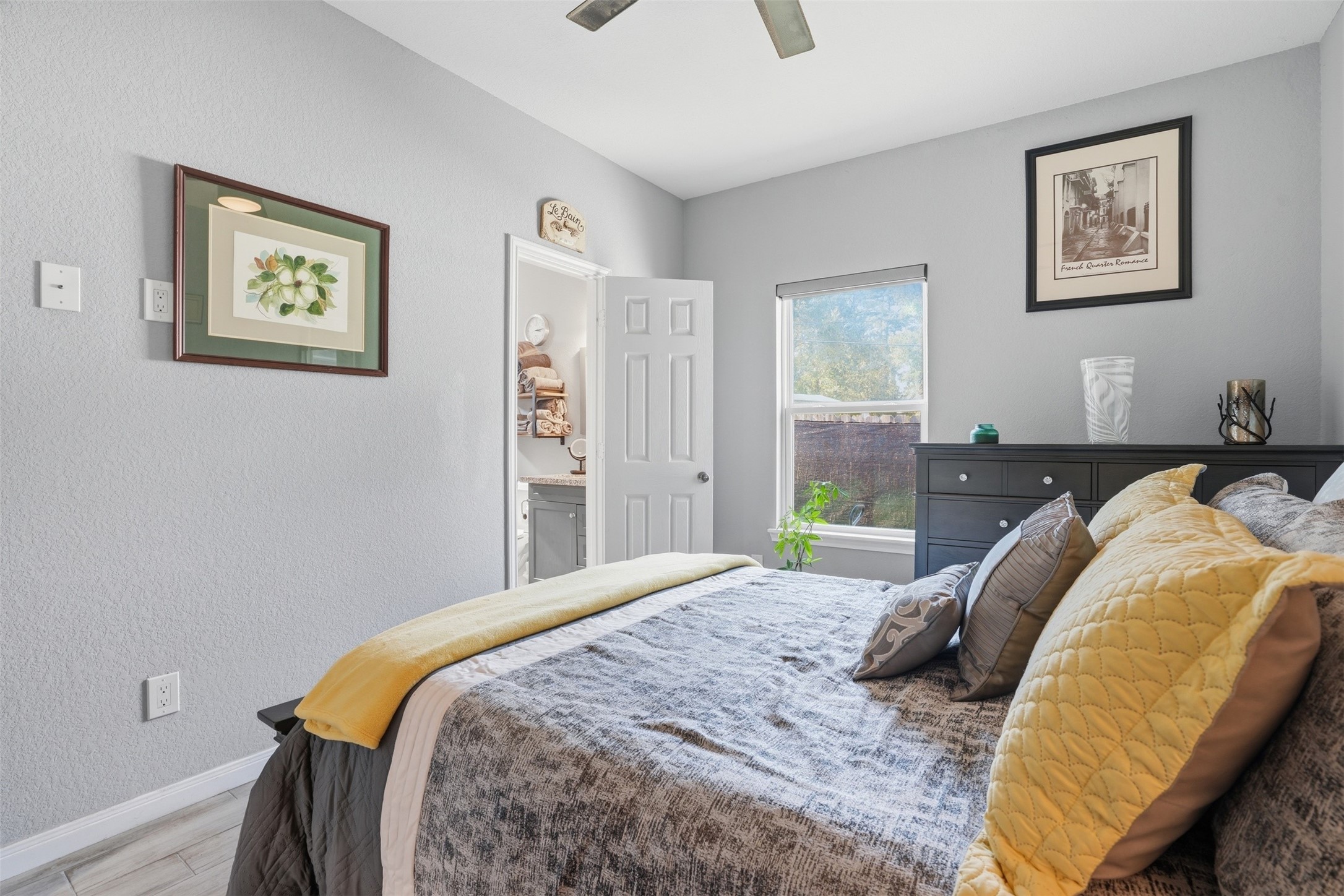 Primary bedroom with soft gray walls, featuring a neatly made bed adorned with colorful pillows, ample natural light from a large window, and direct access to a en-suite bathroom.