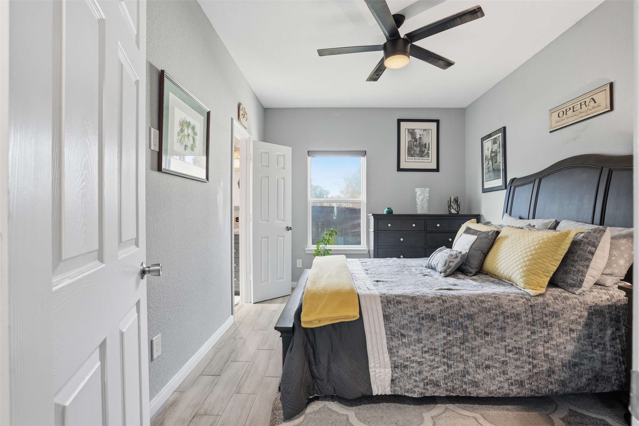 Primary bedroom with soft gray walls, and a large window that allows natural light to fill the space. The room is elegantly decorated with framed artwork and a plush bed adorned with complementary pillows.