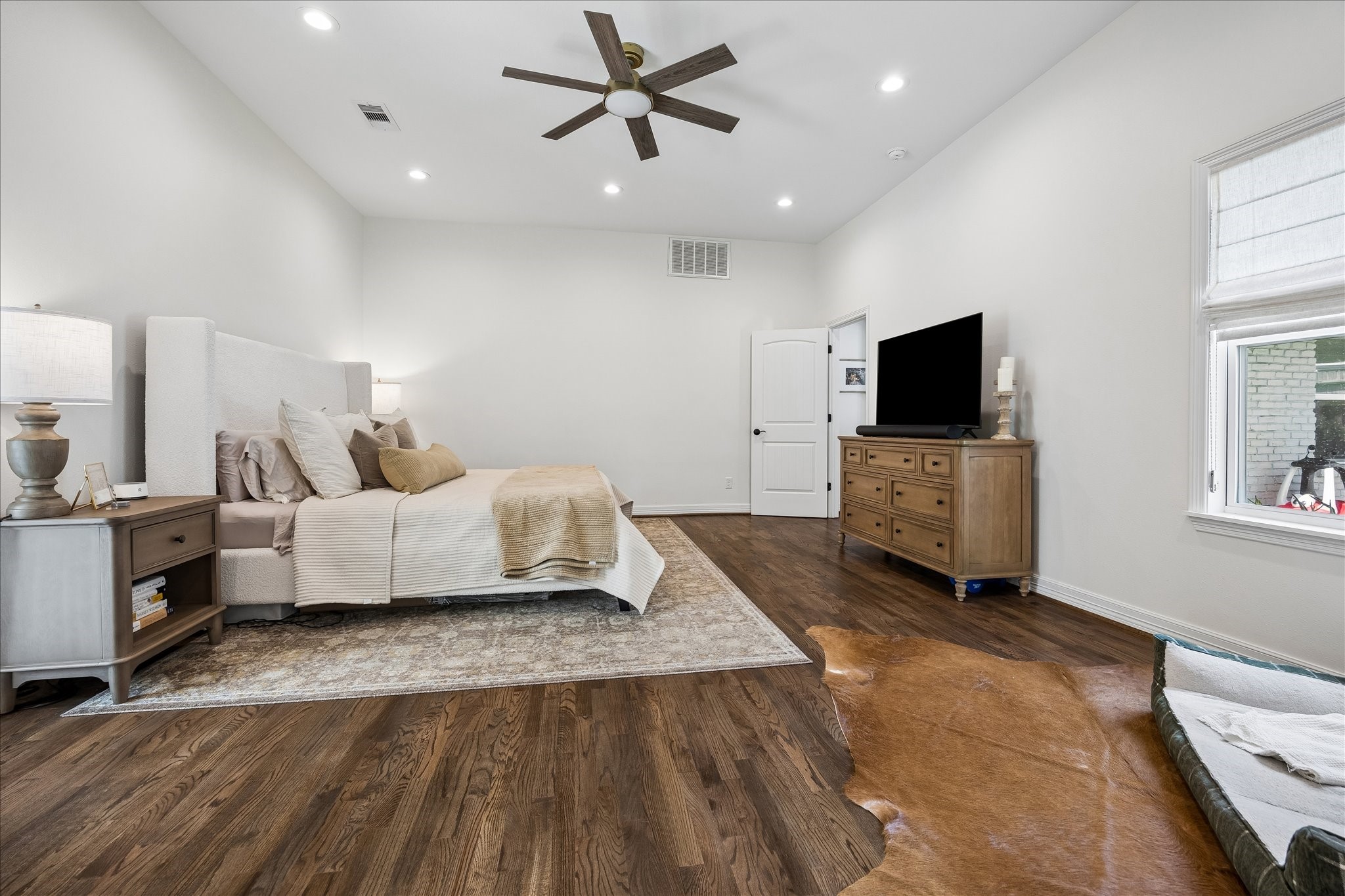 Spacious bedroom featuring a cozy bed with neutral tones, elegant hardwood floors, and a ceiling fan. Natural light streams through the window, enhancing the room's warmth and inviting atmosphere.