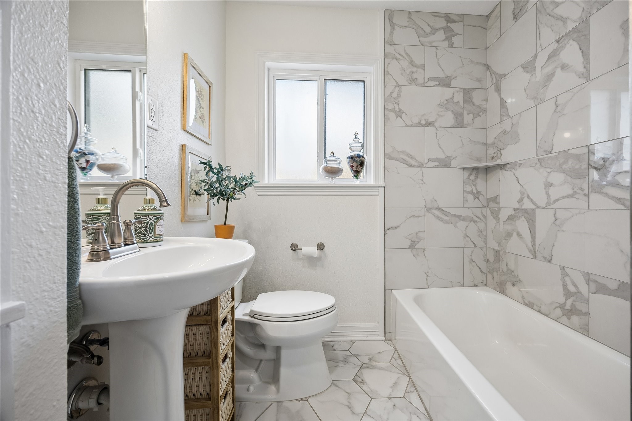 Elegant hall bathroom featuring marble-patterned tiles, a sleek pedestal sink, and a classic bathtub, complemented by modern fixtures and natural light from a frosted window.