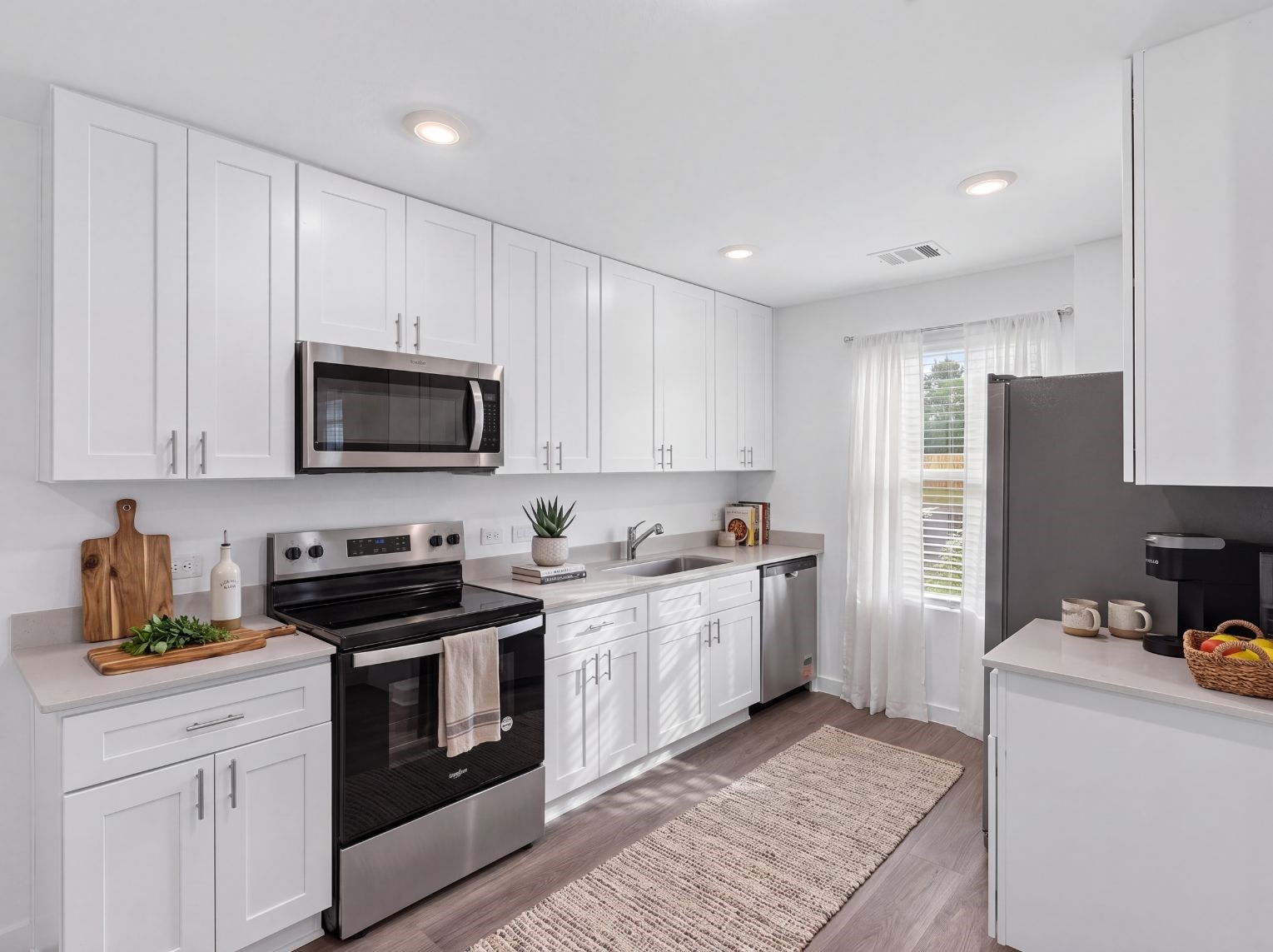 Full view of the kitchen showing ample white cabinetry, stainless steel appliances, light counters, and a window above the sink. Wood-look flooring and a walk-through layout add function.
This photo has been digitally staged to illustrate the possibilities of the home's aesthetic and potential design. Some furnishings and dcor shown are for visual inspiration.