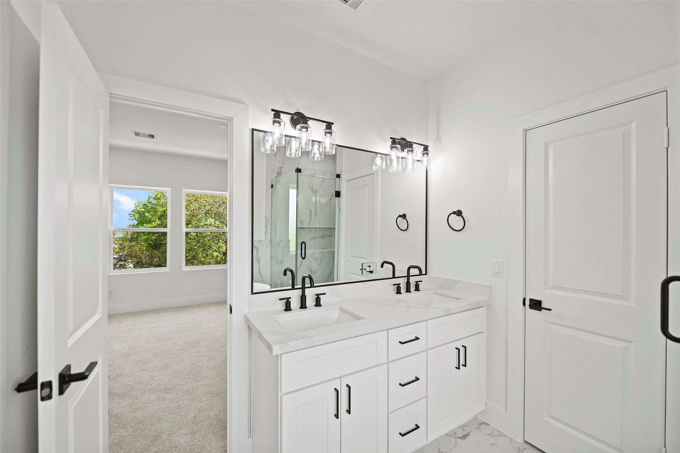 This bright, modern primary bathroom features a double vanity with sleek black fixtures, elegant lighting, and marble like quartz countertops.  It open to the spacious, carpeted primary bedroom with large windows offering a view of lush greenery.