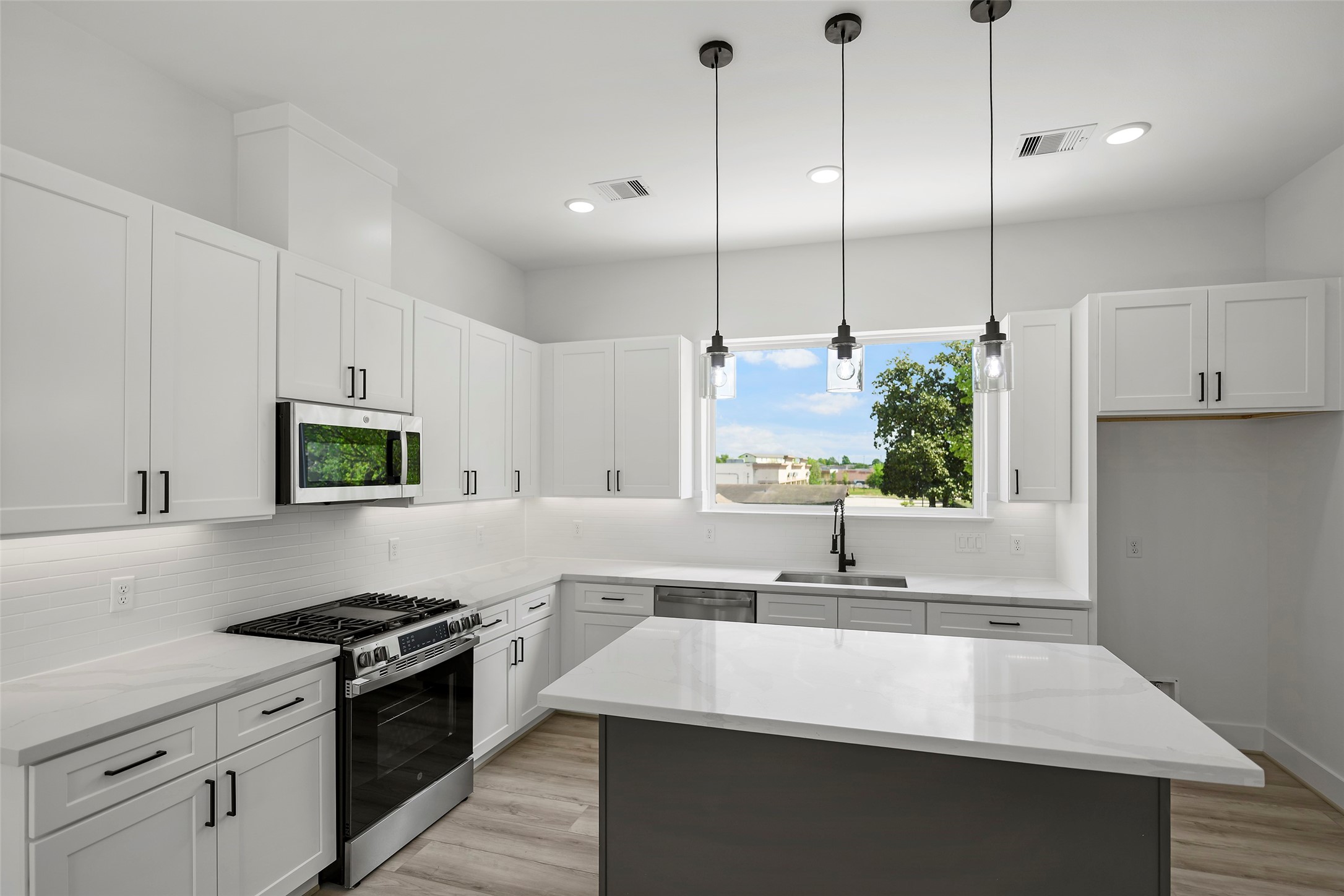 This modern kitchen features sleek white cabinetry, stainless steel appliances, and a spacious island with pendant lighting.  If offers ample natural light through a large window, creating a bright and inviting space.