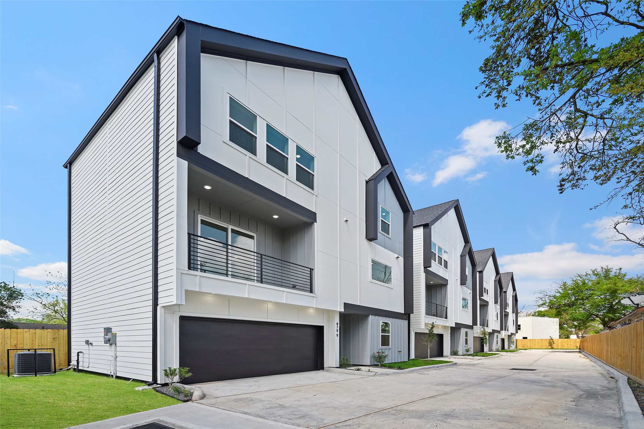 Modern townhouse with a sleek, angular design, featuring multiple stories and a two-car garage.  The exterior is a mix of white and dark accents, with large windows and a small balcony.  situated in a row of similar homes.  It offers a clean, contemporary look in a well-maintained neighborhood.
