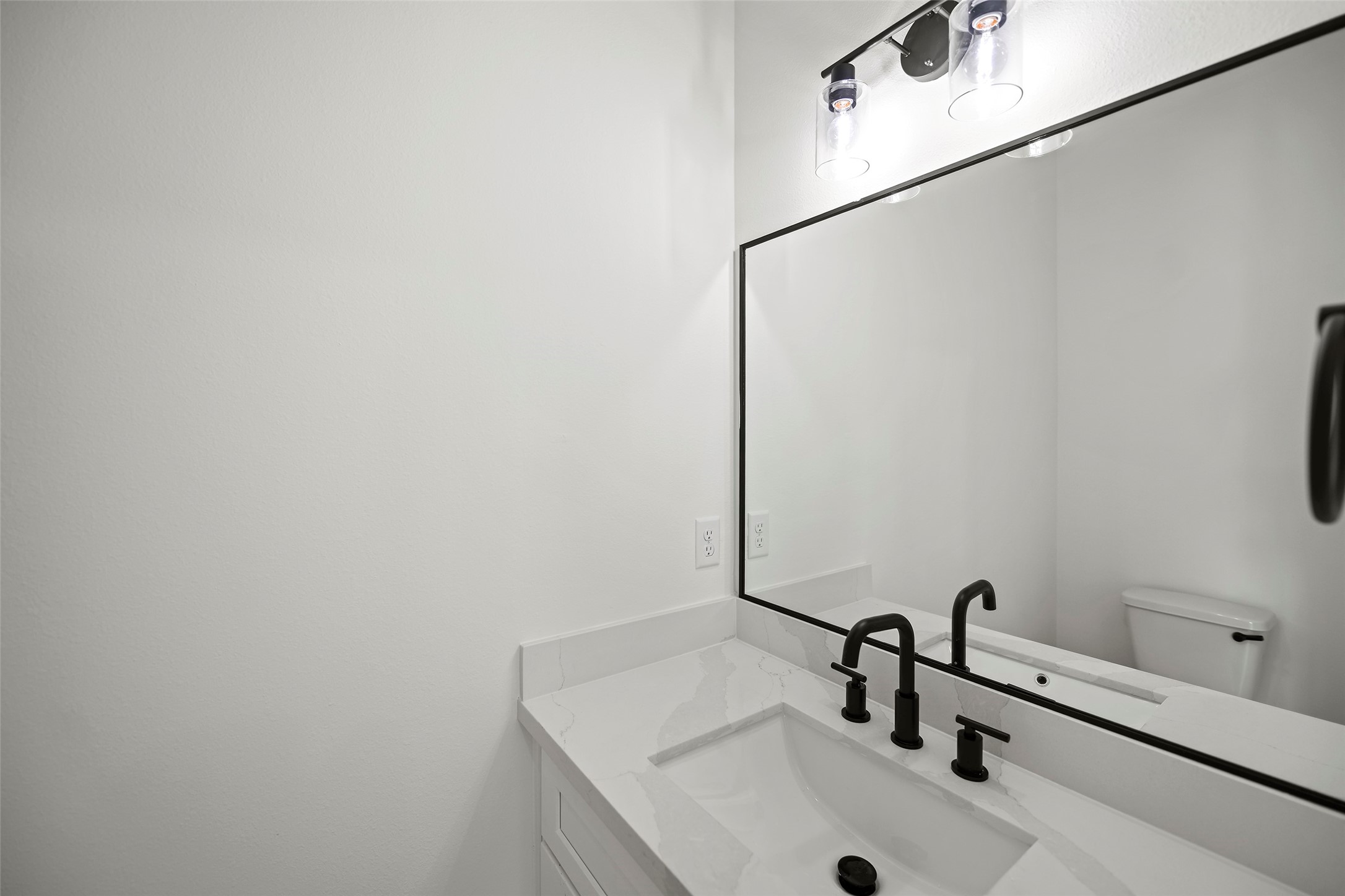 Modern half bathroom with a sleek white vanity, black fixtures, a large mirror, and bright lighting.