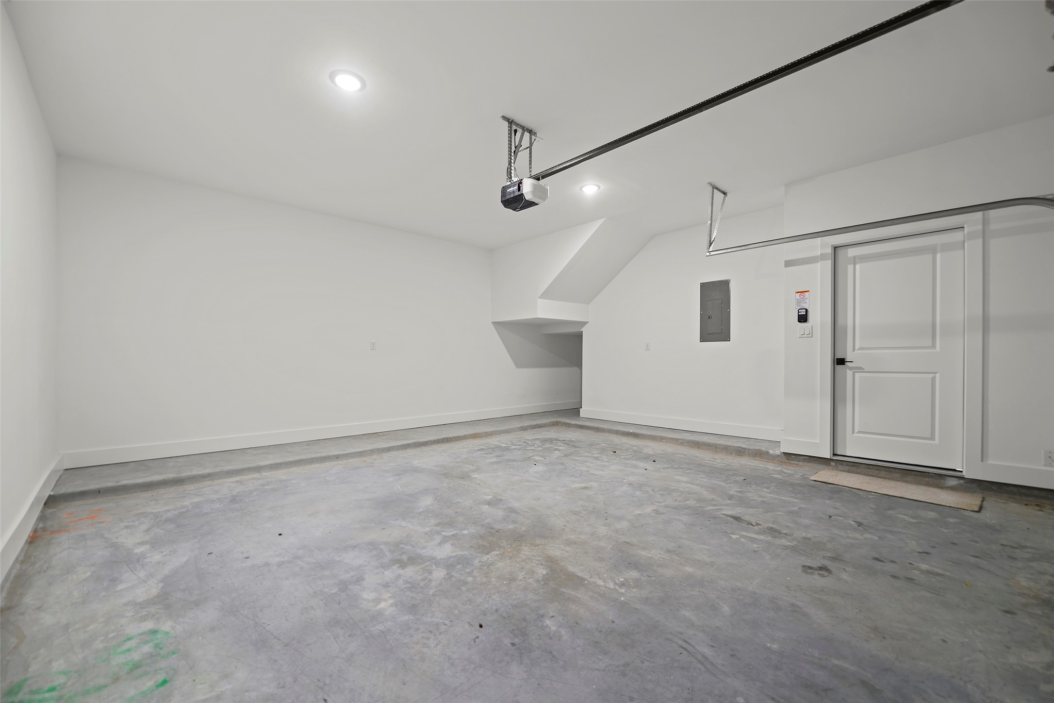 This photo shows a spacious, clean, and well-lit garage with concrete flooring, white walls, and a door leading inside.  It features a garage door opener and ample space for parking or storage.