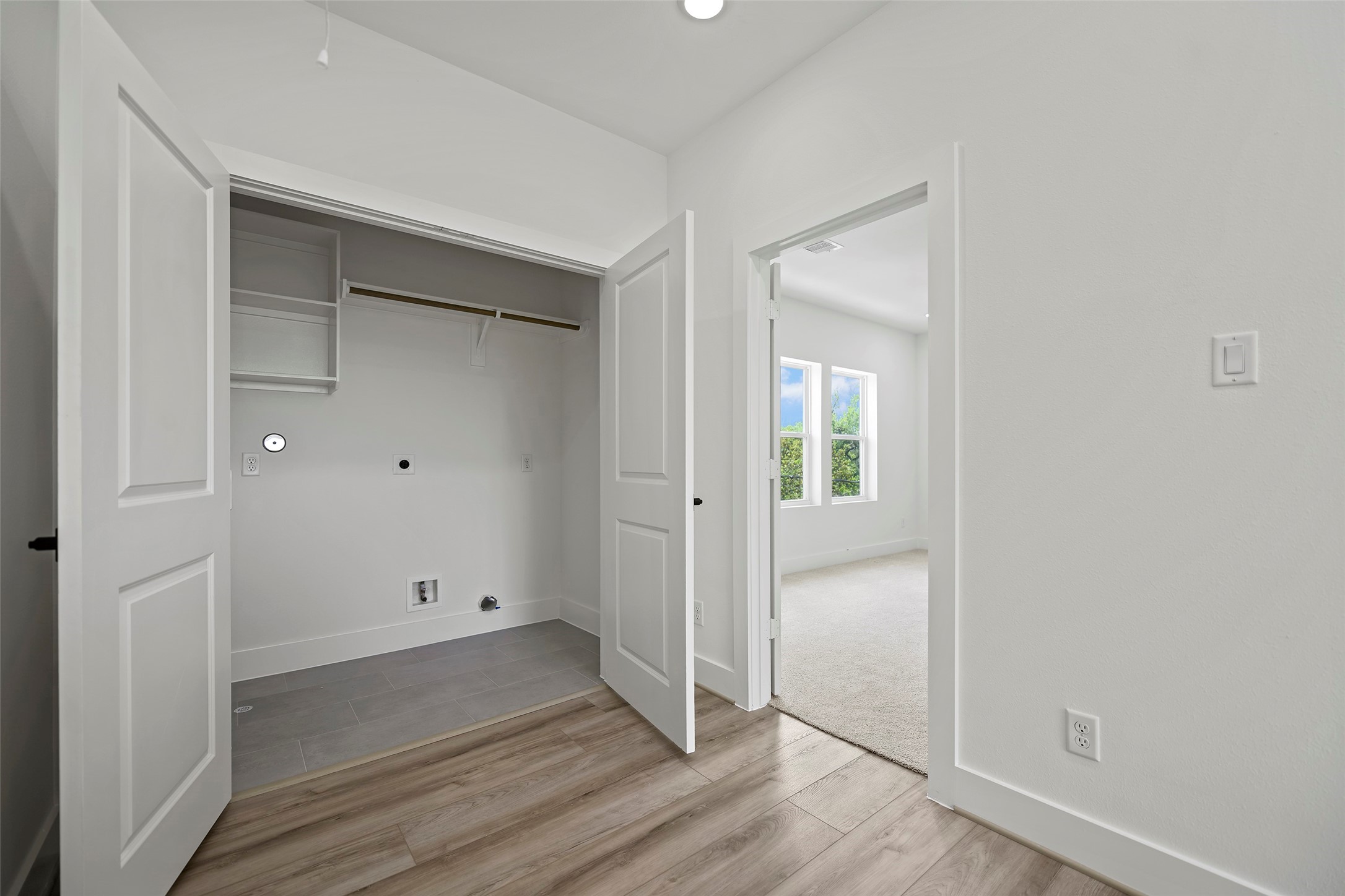 The modern laundry area with double doors, shelving, and space for appliances is located on the 3rd level.  It leads into the primary bedroom with carpeted flooring and large windows, allowing plenty of natural light.  It has a clean, fresh look with neutral tones.