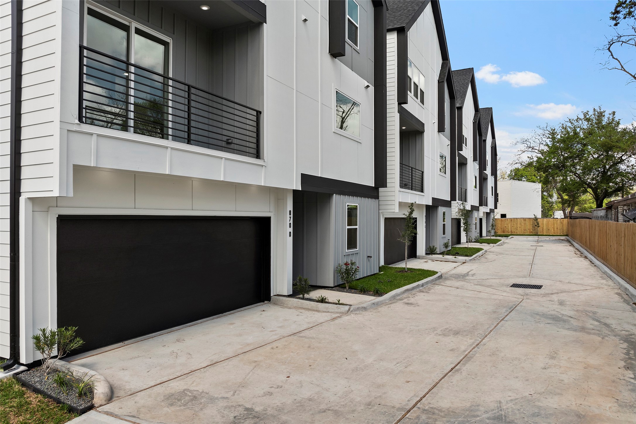 Modern townhomes with sleek black and white exteriors, featuring spacious garages and balconies.  The driveway is wide and well-paved, lined with young trees and a privacy fence.