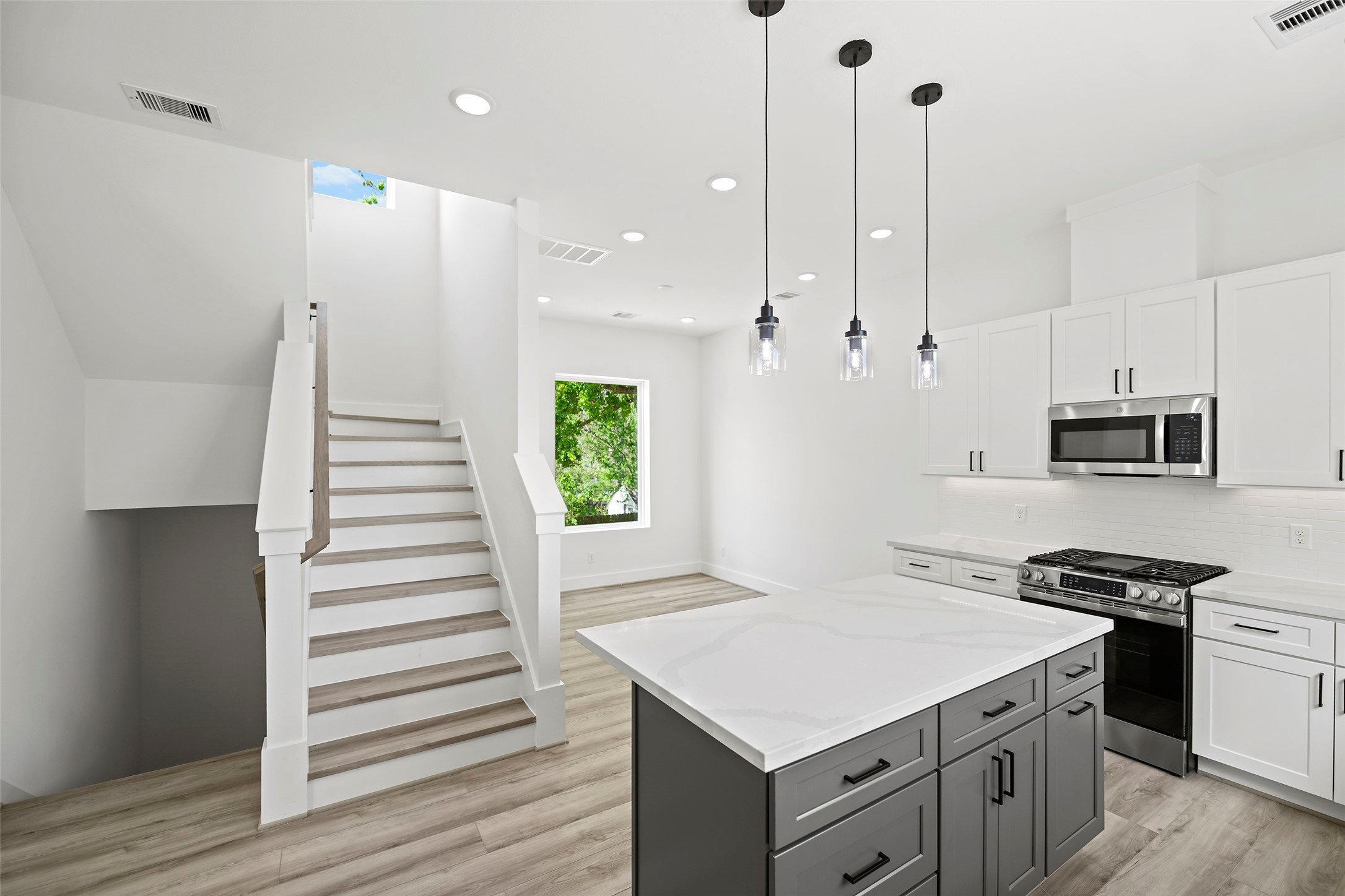 Bright and modern kitchen with sleek white cabinetry, stainless steel appliances, and a spacious island.  The room features elegant pendant lighting and an open staircase, offering a fresh, contemporary feel.
