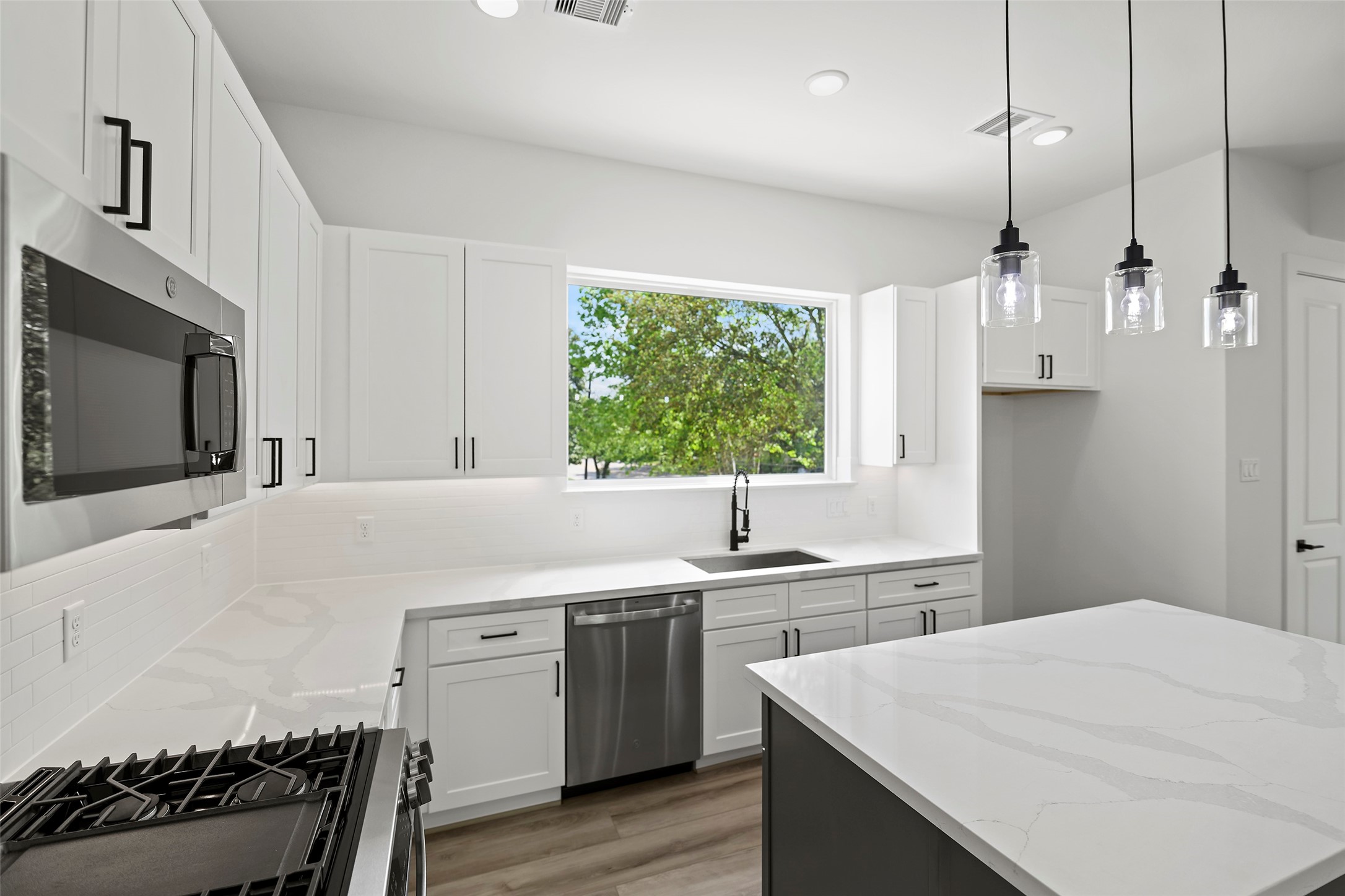 This modern kitchen features sleek white cabinetry, stainless steel appliances, and a spacious island with pendant lighting.  It offers ample natural light through a large window, creating a bright and inviting space.