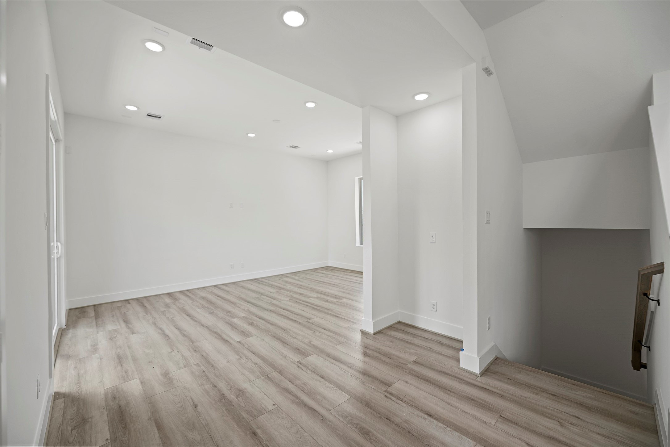The living area showcases a bright and spacious room with modern LVP flooring, recessed lighting, and natural walls.  It features an open layout with a staircase leading down, ideal for versatile use and customization.