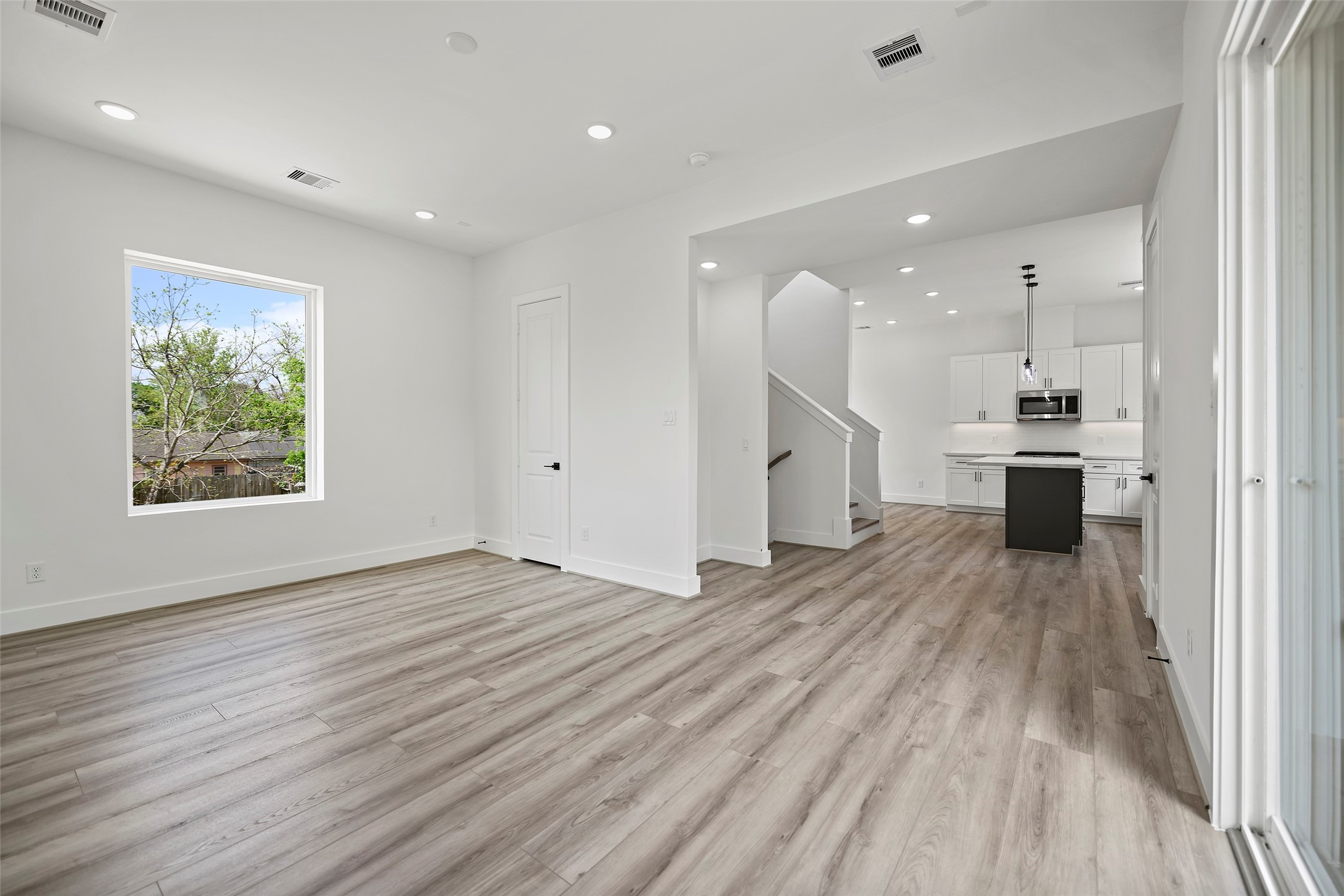 This view from the living area to the kitchen showcases a bright, open-concept living space with modern LVP flooring.  The room flows seamlessly into a sleek kitchen featuring white cabinetry, a central island, and stainless steel appliances.  Large windows provide natural light, and there's a view of the outdoors.