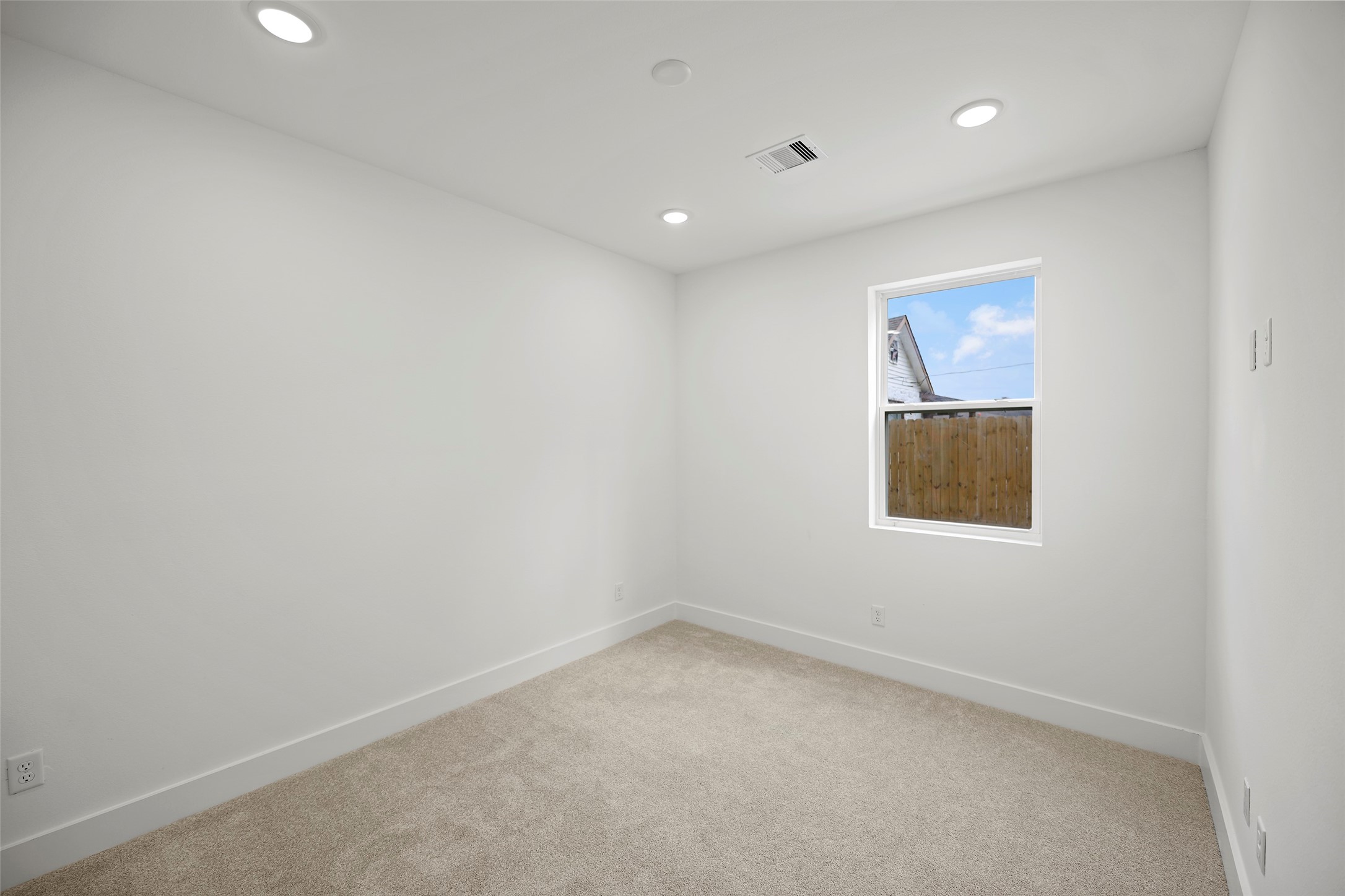 This first level bedroom features a clean, modern design with white walls and recessed lighting.  A single window allows natural light, complemented by plush carpeting, creating a cozy and inviting space.