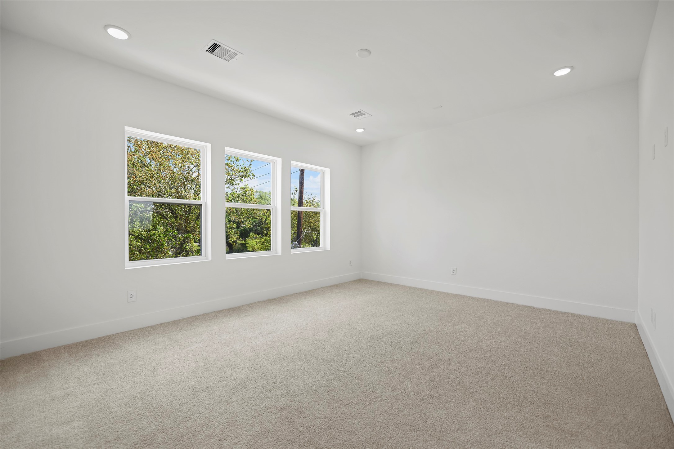 This bright and spacious Primary Bedroom is located on the 3rd level.  It has neutral carpeting and white walls.  It features three large windows bringing in natural light and has recessed lighting in the ceiling.