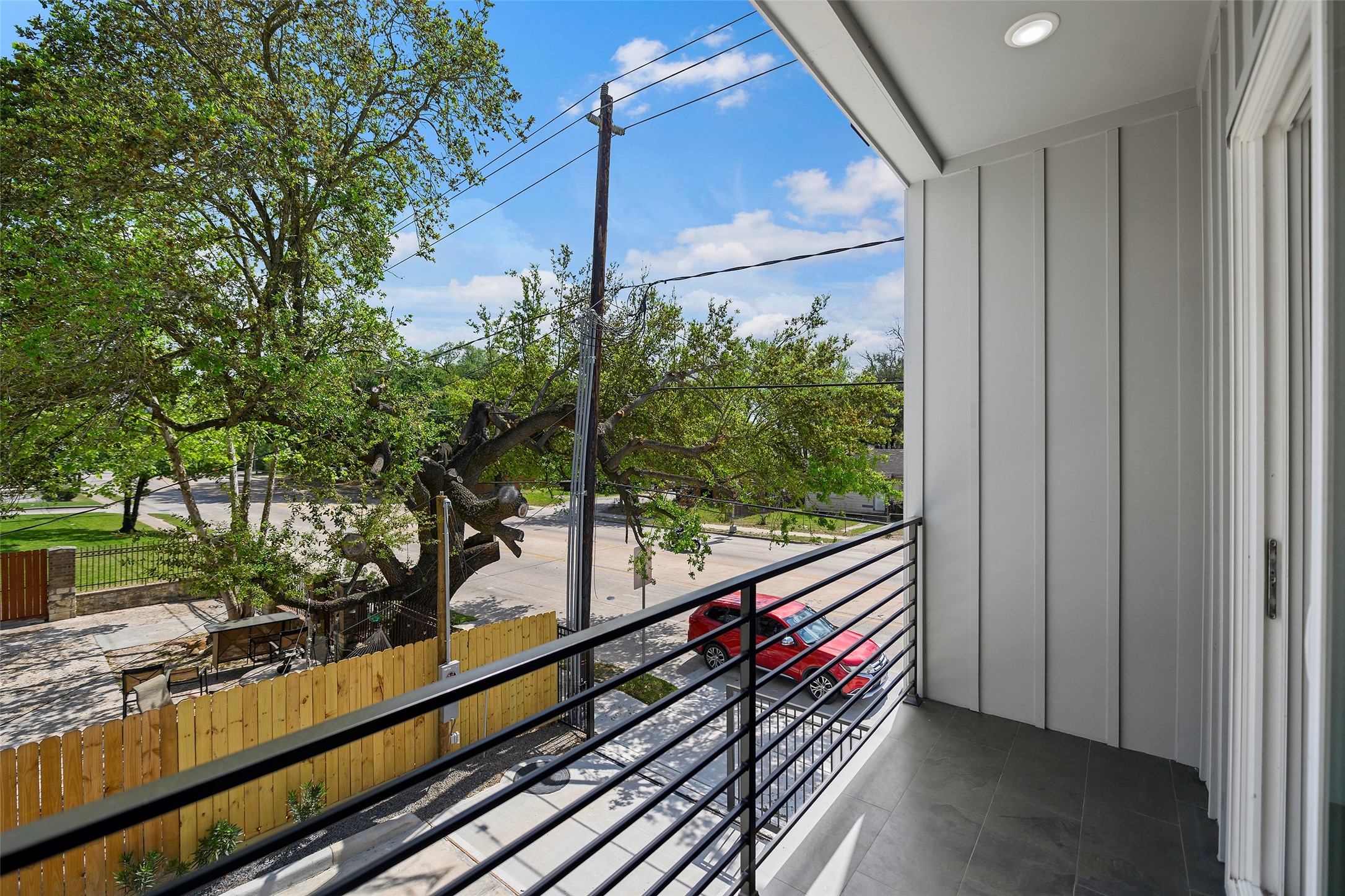 The modern balcony has a sleek metal railing, overlooking a tree-lined street and a wooden fence.  The area is bright with natural light, offering a pleasant view of the neighborhood.