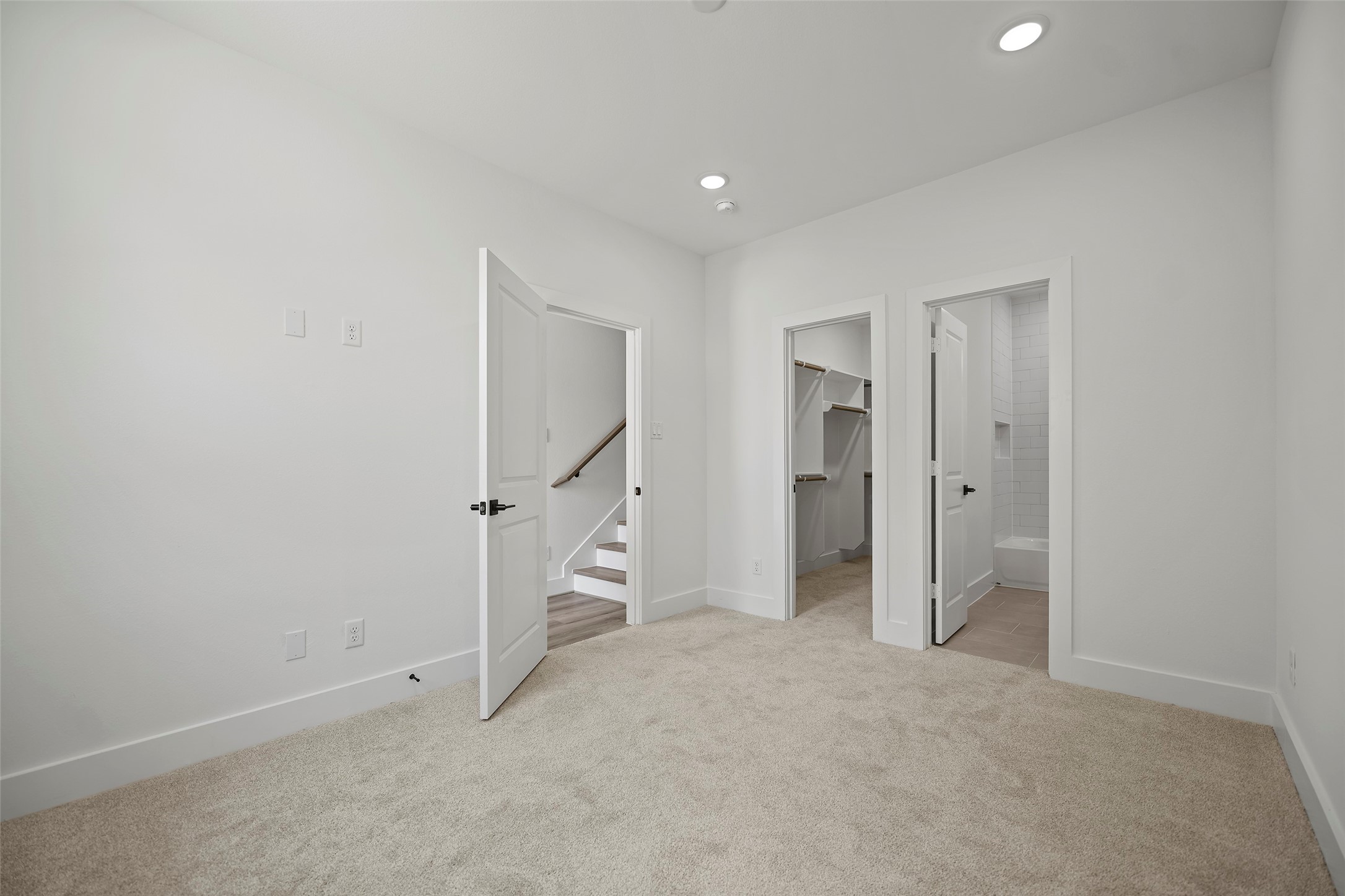 This first level bedroom shows a bright, carpeted room with neutral color palette.  It features three doors:  one leading to stairs, another to a spacious walk-in closet, and the third to a modern bathroom with tile flooring.  Recessed lighting adds a contemporary touch.