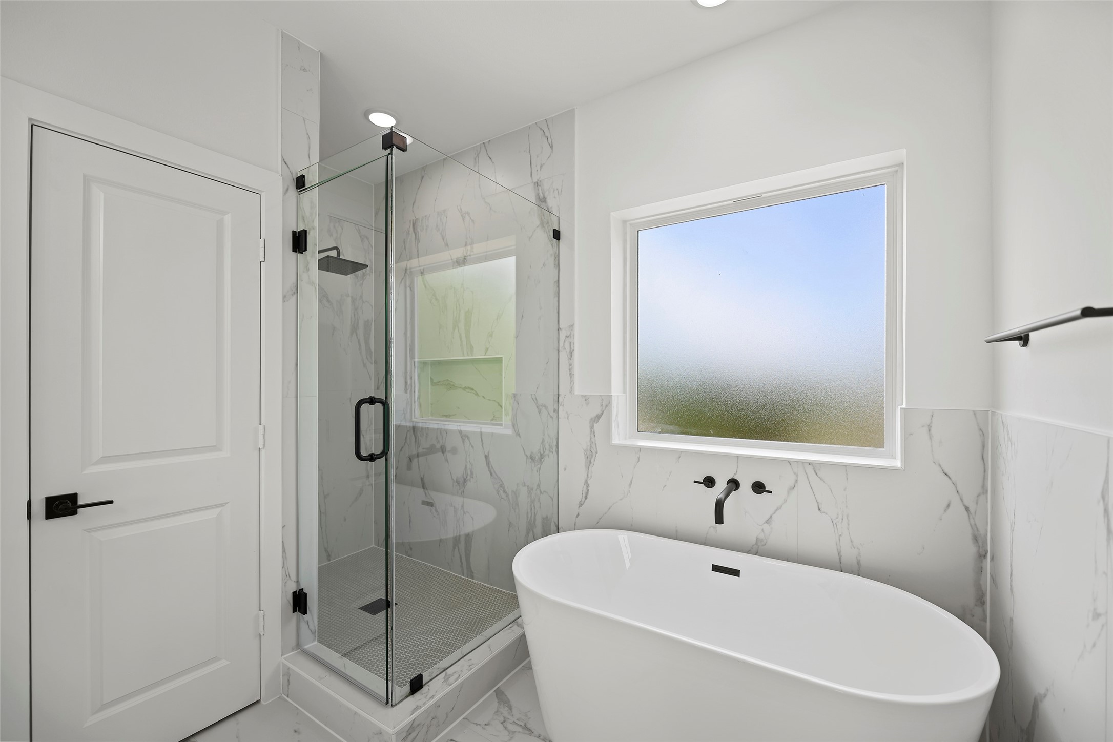 This modern primary bathroom features a sleek freestanding tub, a glass enclosed shower, and elegant marble-patterned tiles.  The large frosted window allows natural light while ensuring privacy.
