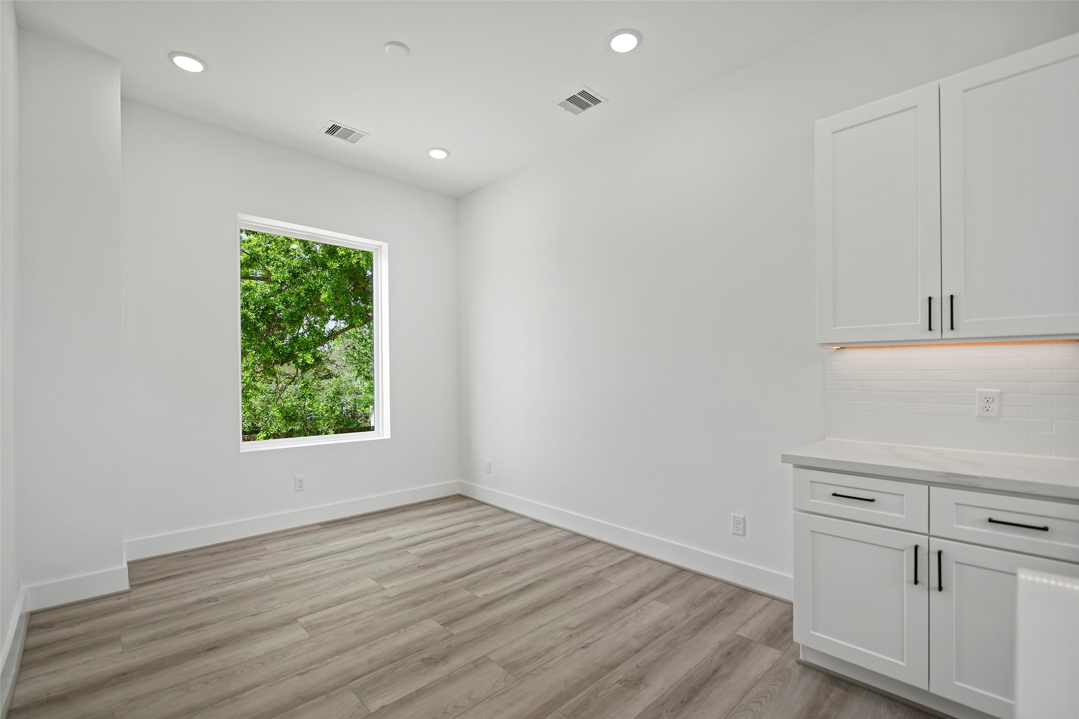The dining area is bright and airy with LVP flooring, recessed lighting, and a large window offering a view of greenery.  Features white soft closing cabinetry and a sleek quartz countertop, creating a clean and modern look.