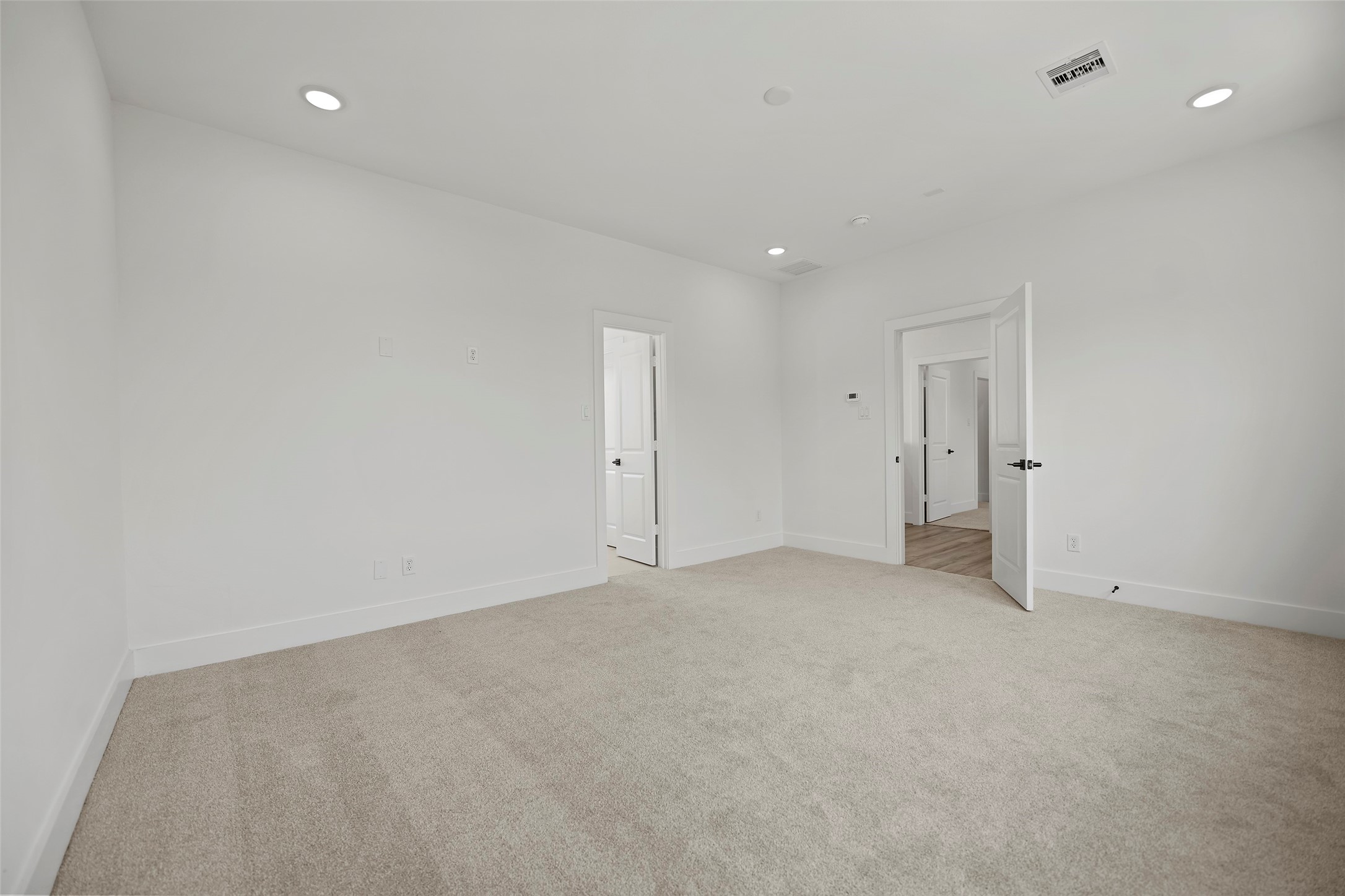 This primary bedroom is bright, spacious and has neutral carpet and walls, featuring recessed lighting and a door leading to the bathroom and another door leading to the adjoining bedroom.