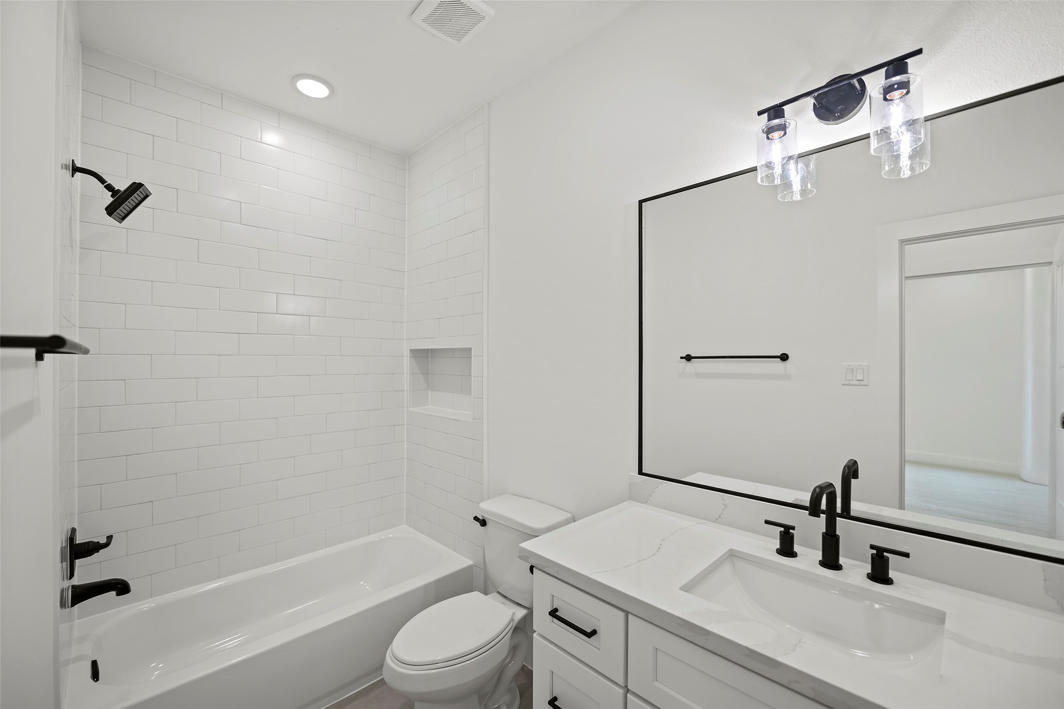 This modern bathroom features a sleek design with a white subway-tiled shower and a built-in niche.  It includes a bathtub, a stylish vanity with a large mirror, and black fixtures for a contemporary look.  The room is well-lit with recessed lighting and decorative light fixtures.
