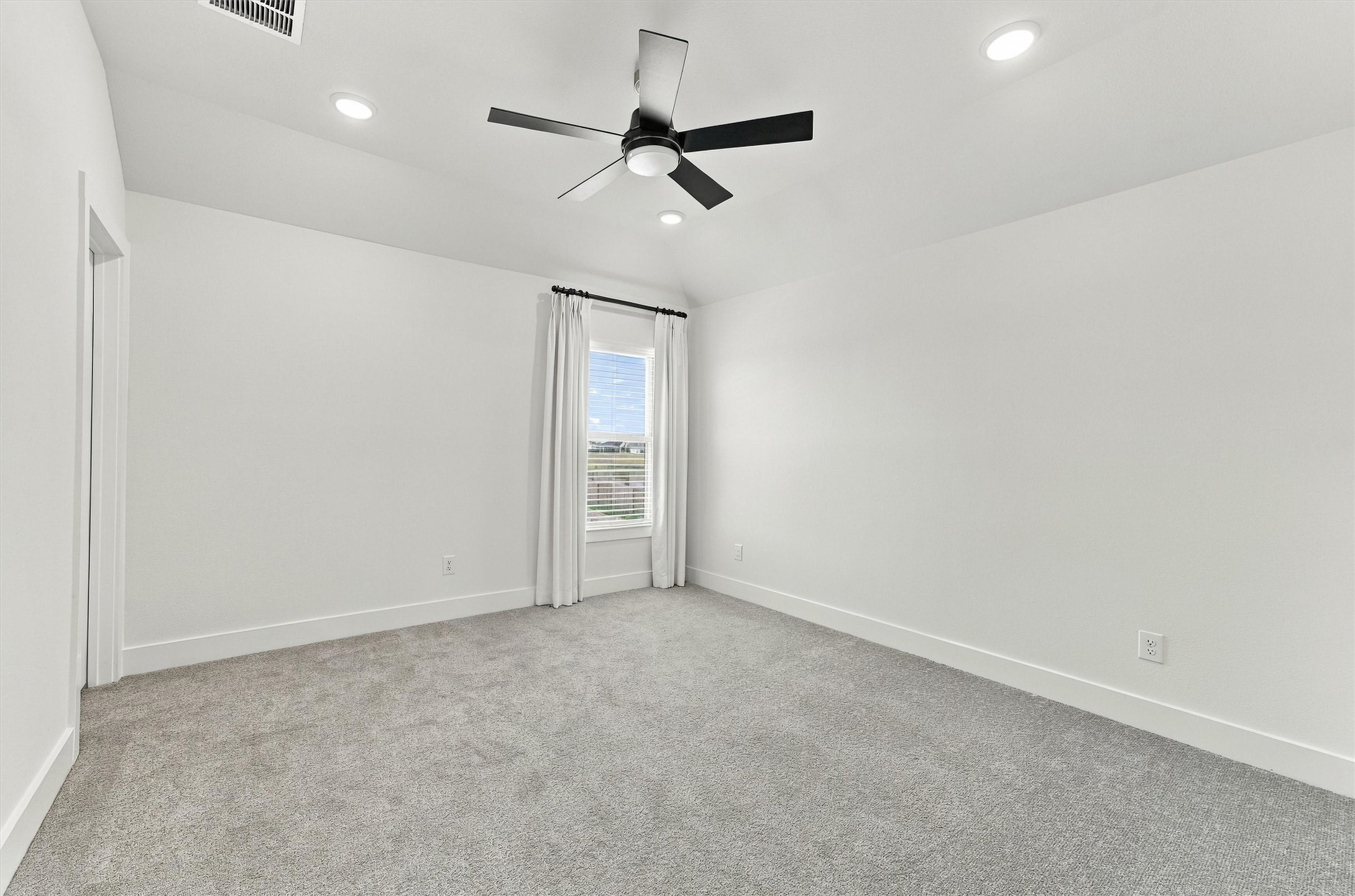A bright secondary bedroom with a ceiling fan, recessed lighting, plush carpet, and tailored drapery.