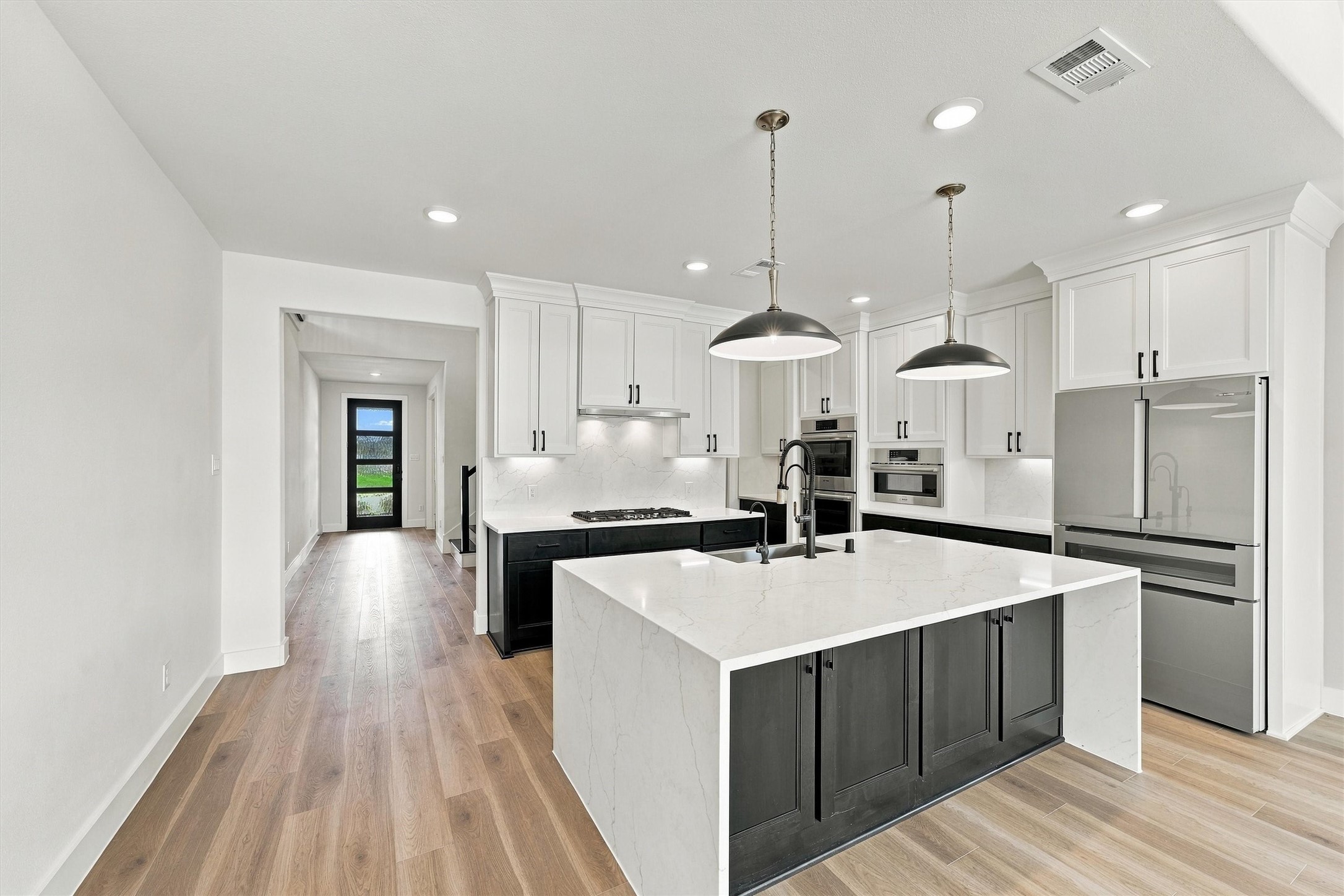 A sculptural waterfall quartz island, gas cooktop, double wall ovens, and two-tone cabinetry, with under-cabinet lighting along a full-height backsplash.