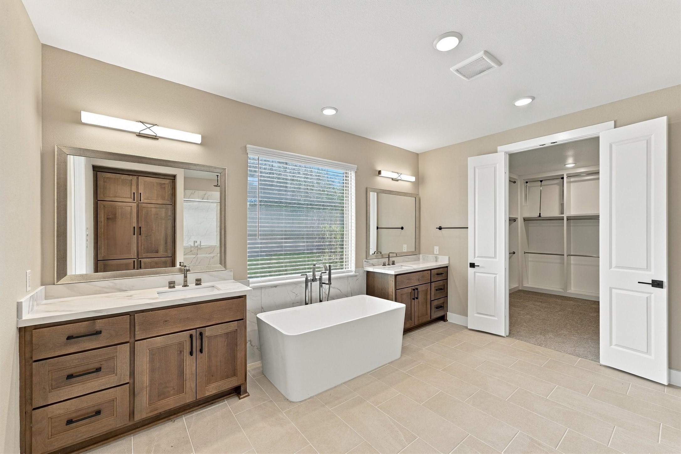 A freestanding soaking tub sits beneath a picture window between separate vanities, with double doors opening to the closet.