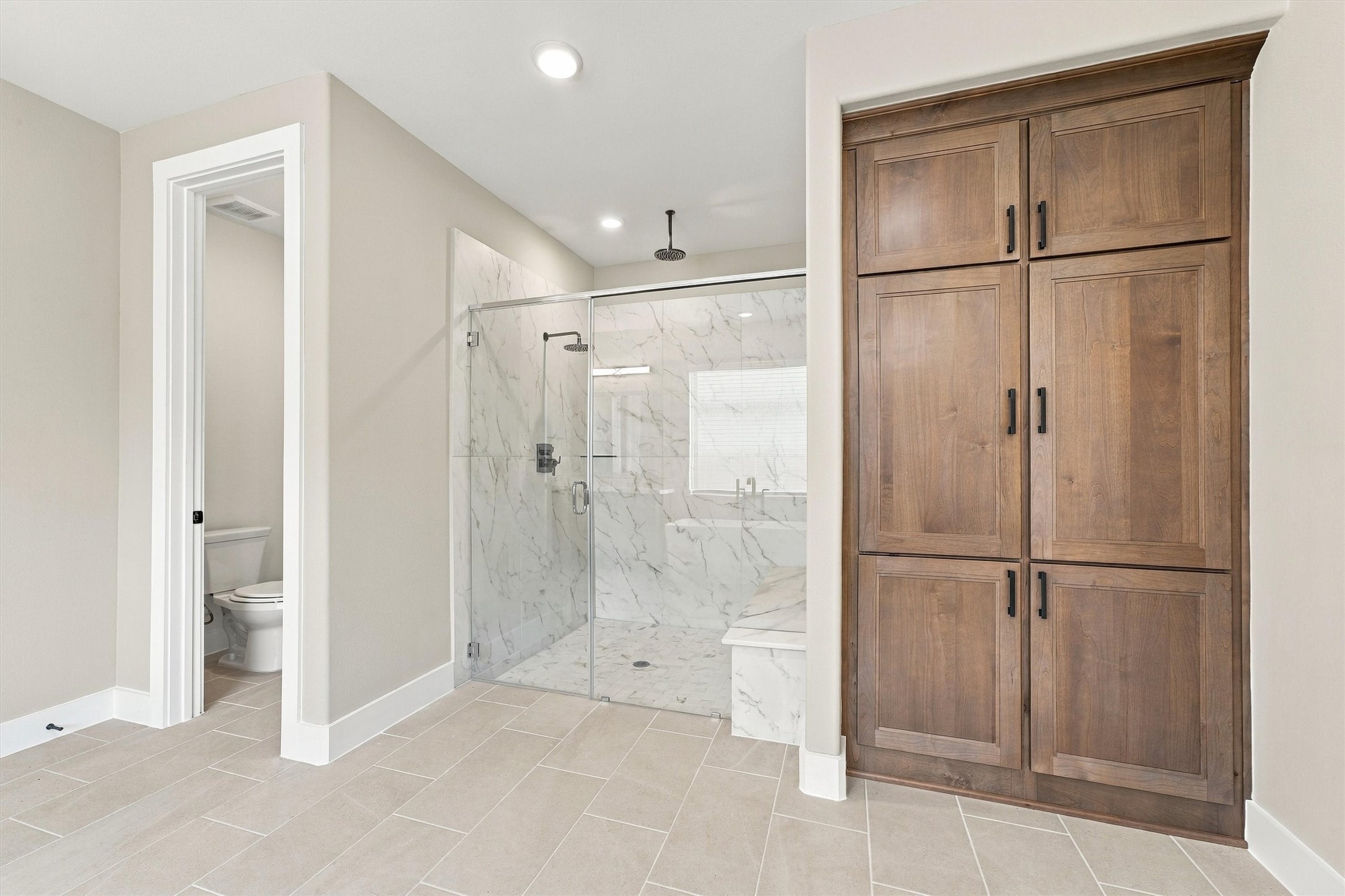 A frameless glass shower with rain head, tile walls, and a built-in bench, next to a custom linen cabinet and a private water closet.