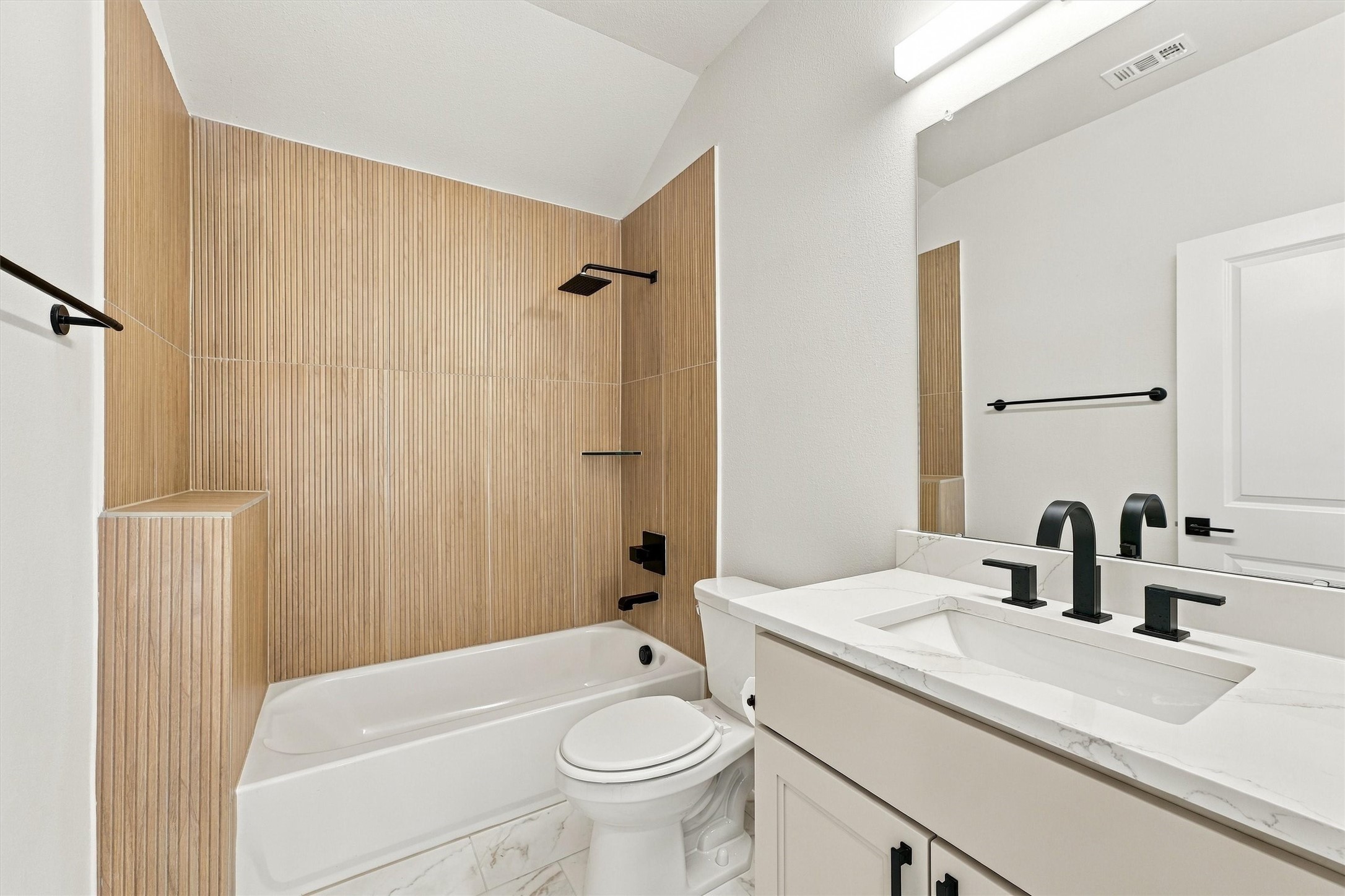 Warm fluted wood-look tile surrounds the tub and shower, with a rain showerhead, matte black fixtures, and a quartz vanity.