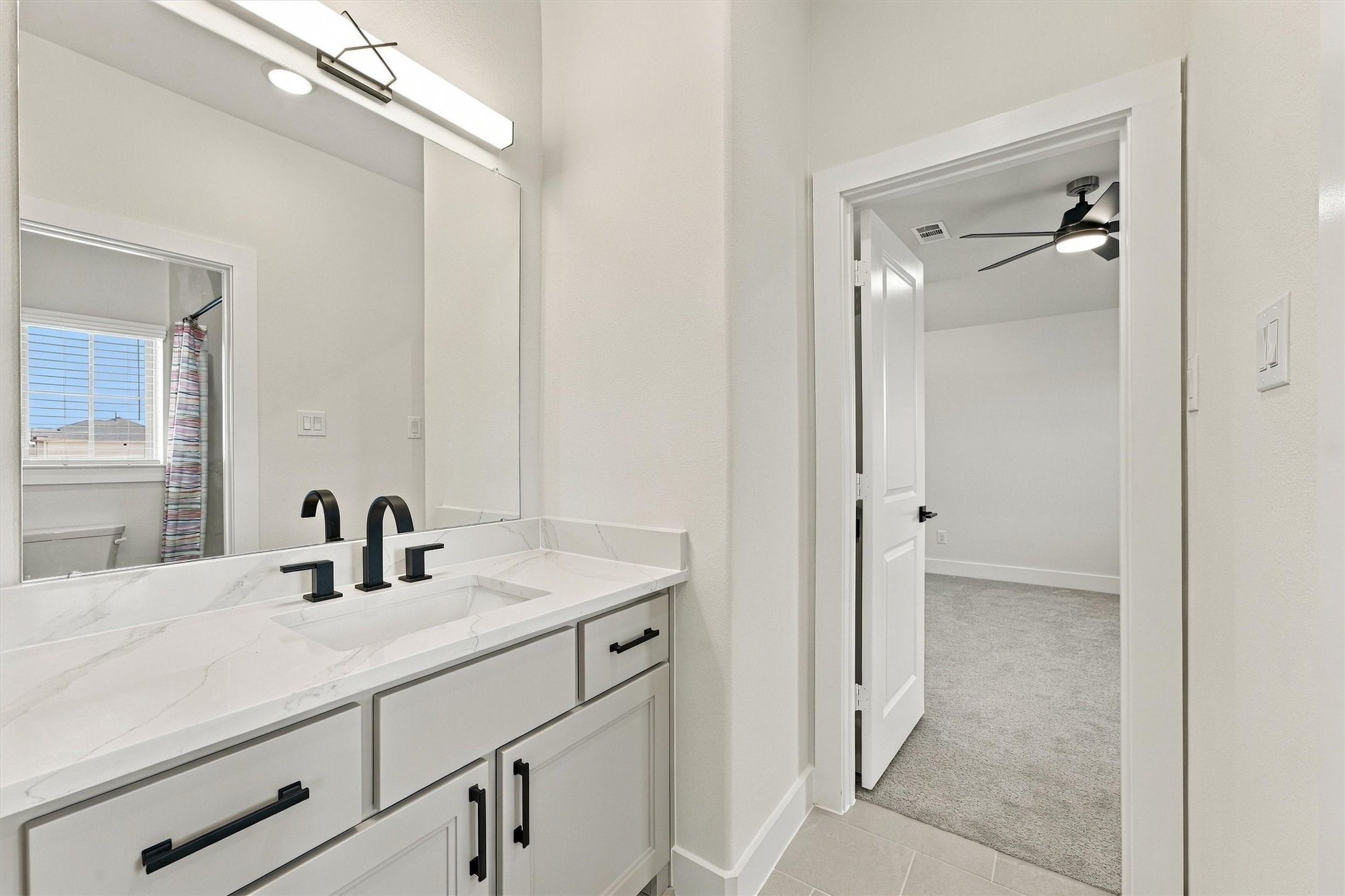 This bath has a quartz vanity, matte black hardware, and a modern light bar, and it opens directly to a bedroom.