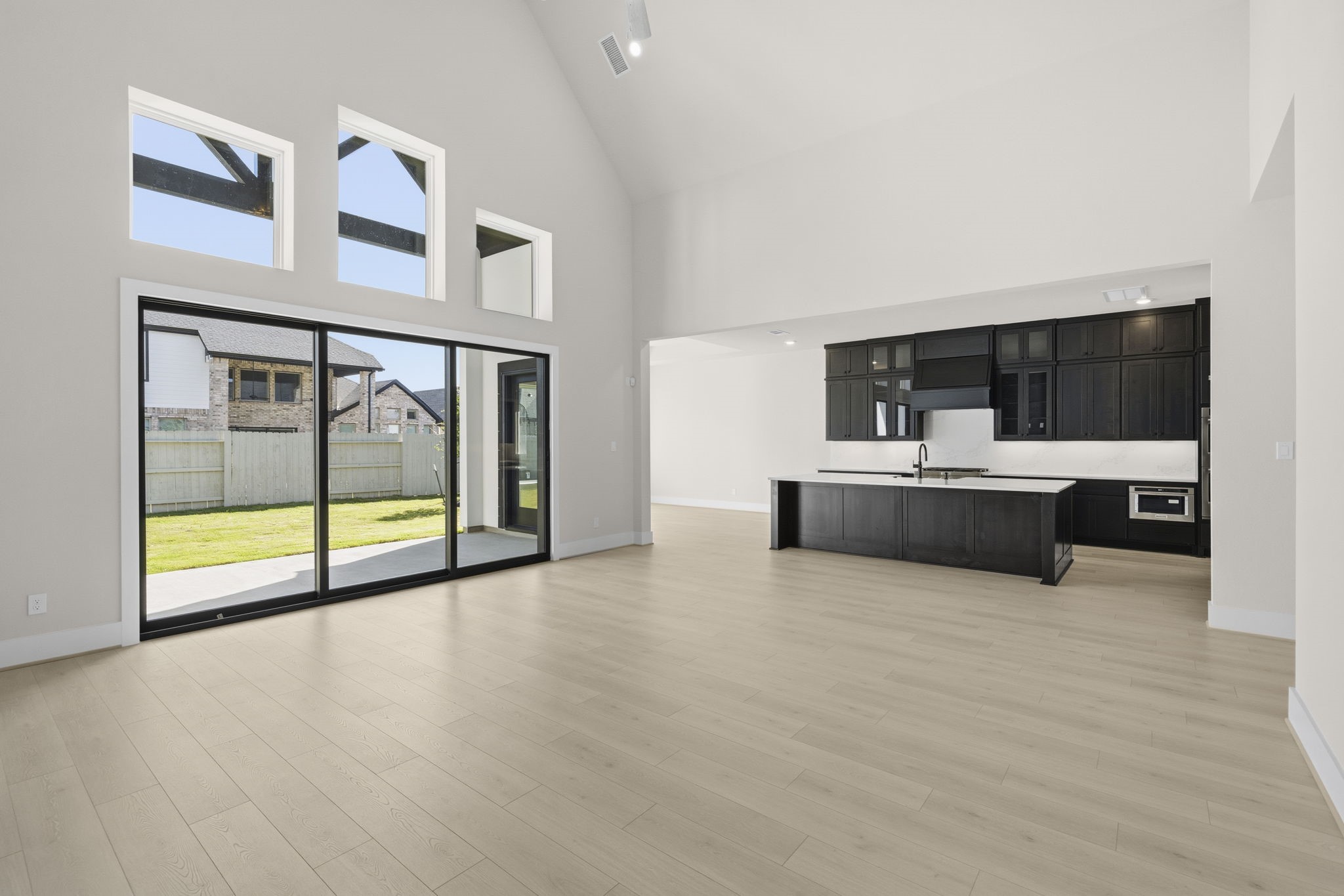 This modern open-concept living space features high ceilings, ample natural light from large windows, and a sleek kitchen with dark cabinetry. The sliding glass doors lead to a spacious backyard, creating an inviting indoor-outdoor flow.