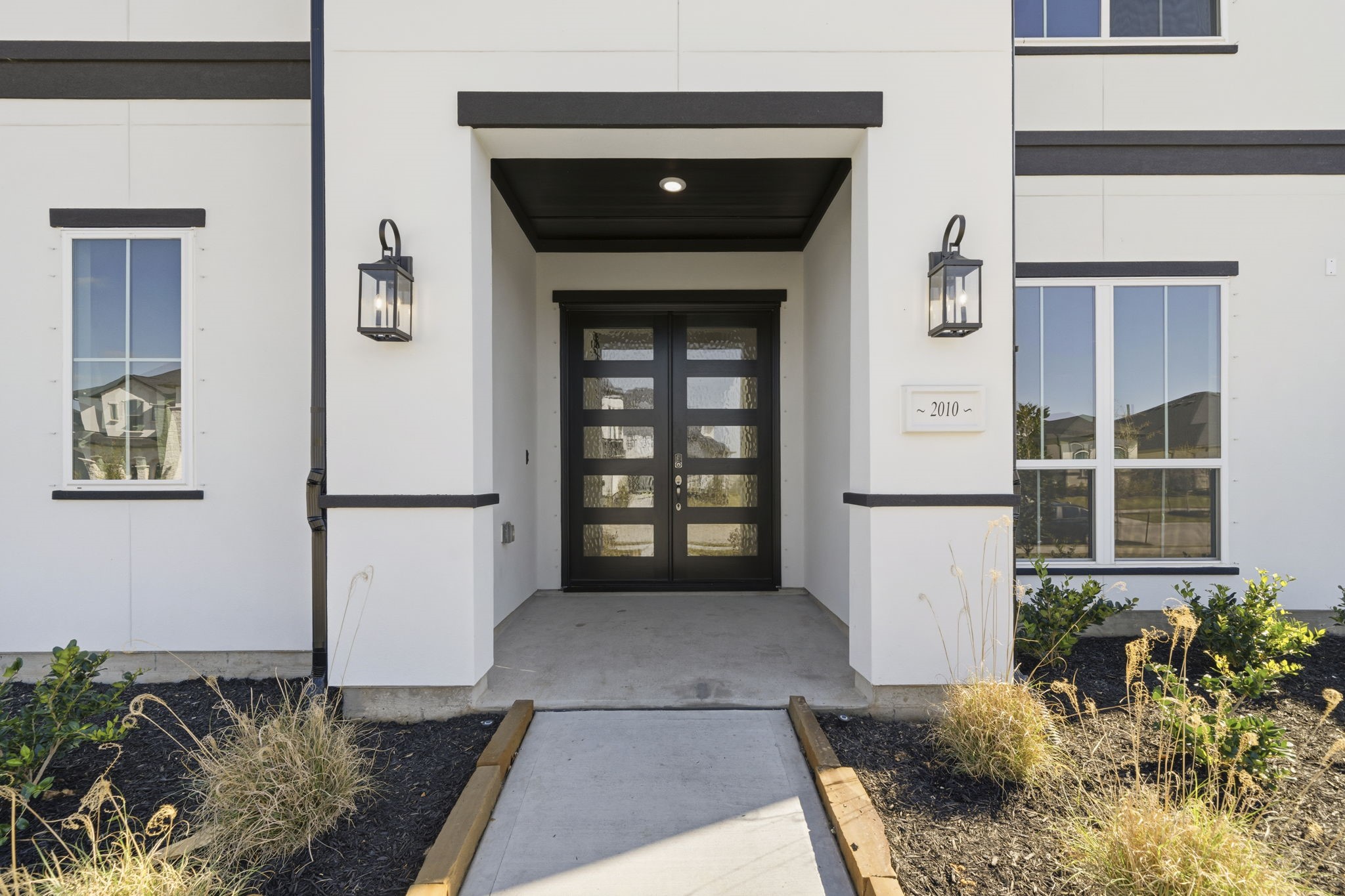 Modern entryway with a black-framed glass door, sleek exterior lighting, and clean landscaping leading up to the entrance.