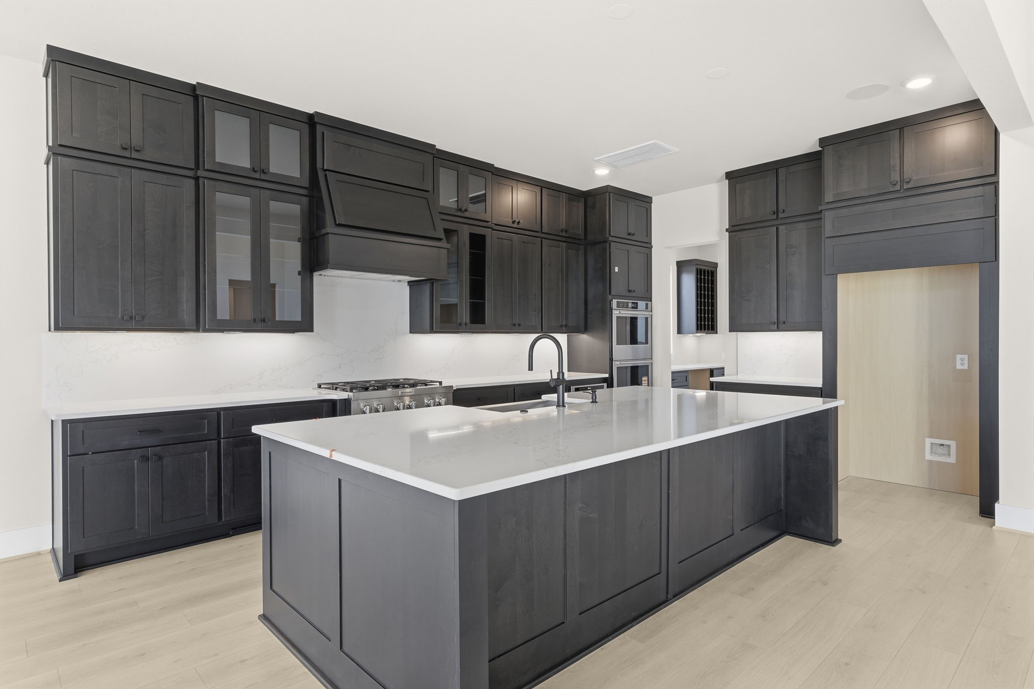 This kitchen features sleek, dark cabinetry with ample storage, a large island with a modern sink, and stainless steel appliances. The light wood flooring and neutral walls create a bright, contemporary space perfect for cooking and entertaining.