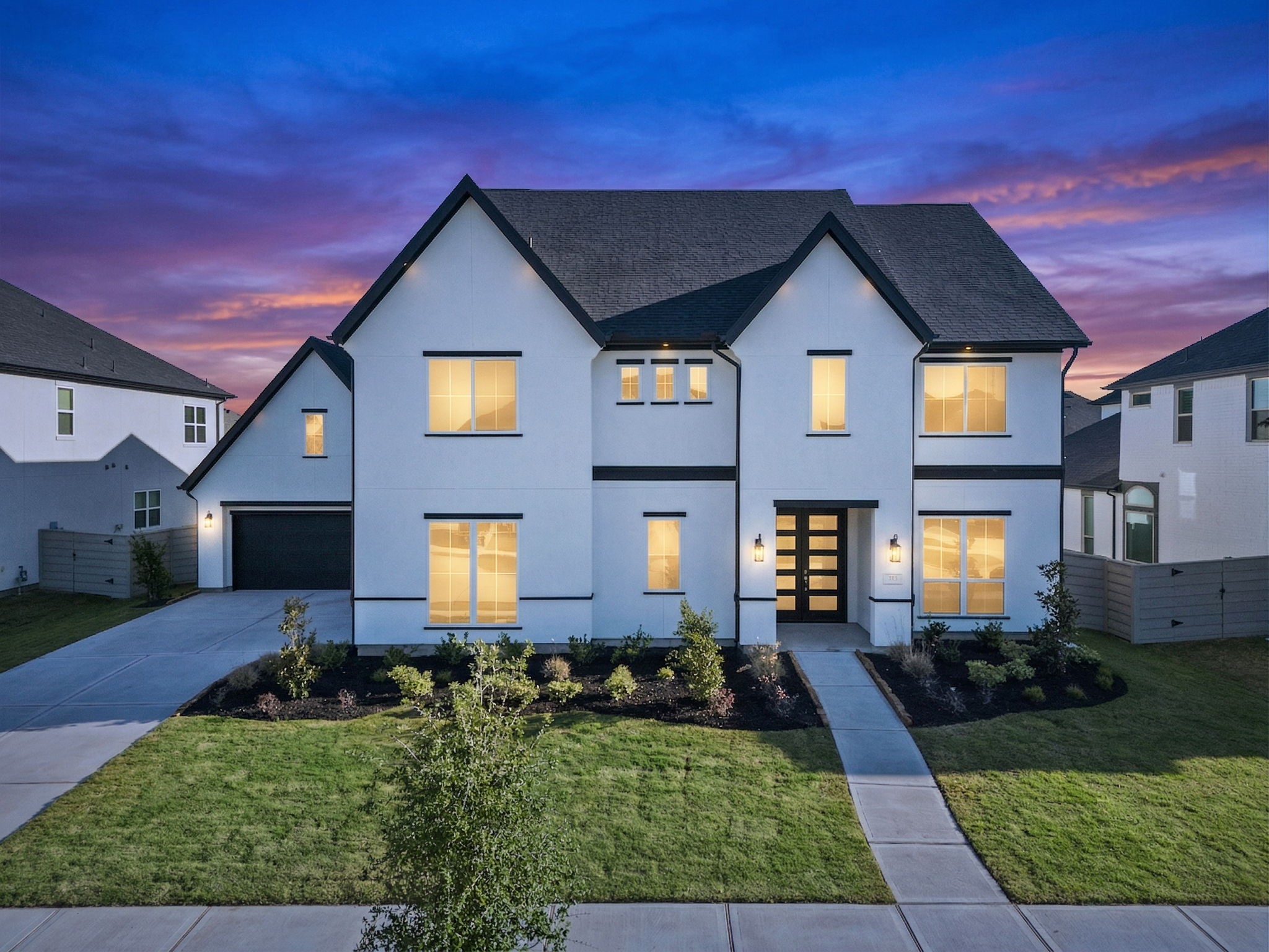 Modern two-story home with a sleek white exterior and dark roof accents. Large windows provide ample natural light, and the well-maintained landscaping enhances curb appeal. The driveway leads to an attached garage, set against a picturesque sunset backdrop.