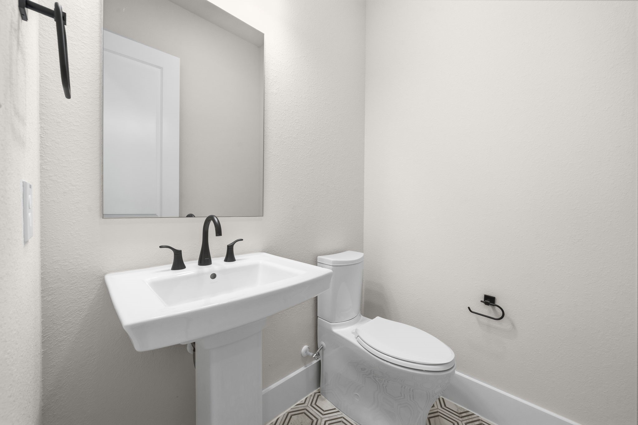 Modern half-bathroom featuring a pedestal sink with sleek black fixtures, a large mirror, and a toilet. The room is bright with neutral walls and patterned floor tiles.