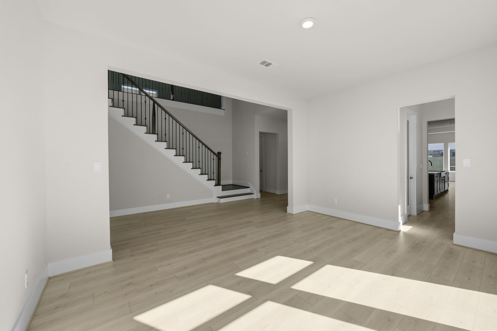 This spacious living area features light wood flooring, neutral walls, and ample natural light. It has an open view of a staircase with elegant railing, and leads into the kitchen, creating a seamless flow.